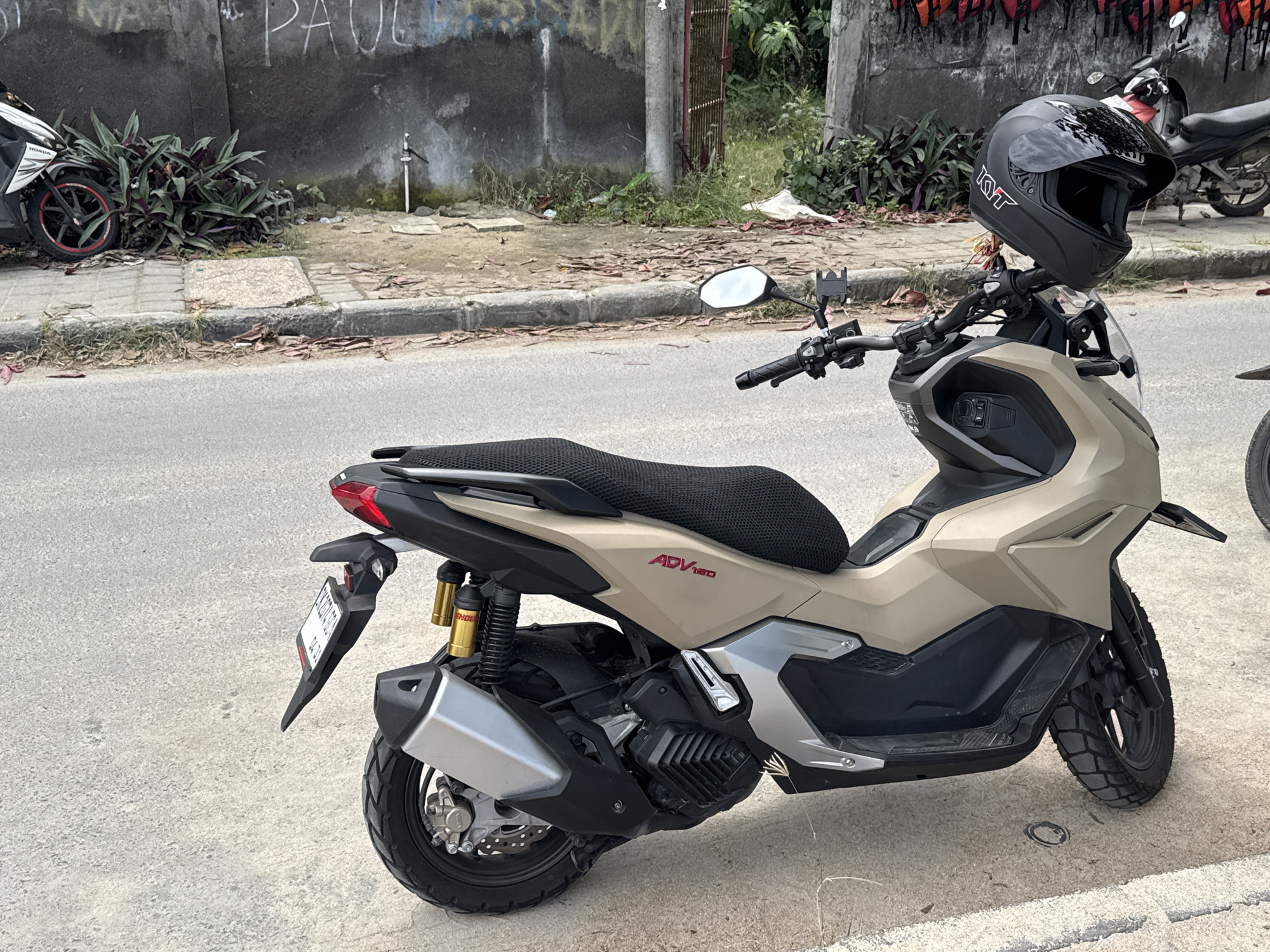 A tan Honda ADV150 scooter is parked on the side of a road in Manggis, Indonesia. A black KYT helmet is perched on the handlebars. The scooter has a black textured seat, a license plate, and gold-colored shock absorbers. In the background, a white Honda scooter with a red rim is parked near a wall with graffiti. The environment includes a sidewalk, vegetation, and a street.