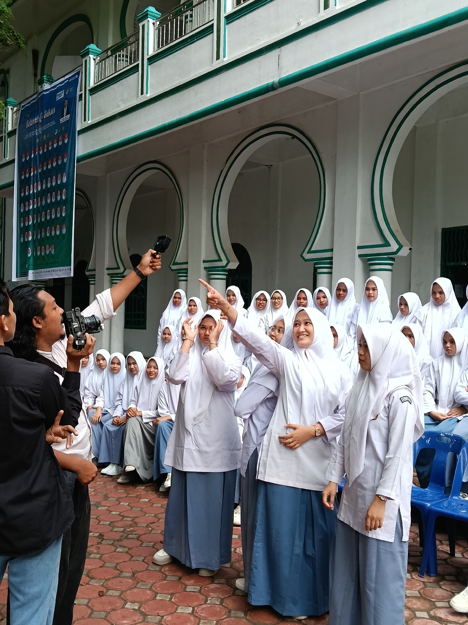 This outdoor image captures a lively scene featuring a group of young women, likely students, dressed in white hijabs, white long-sleeved tops, and long blue skirts or trousers, indicative of school uniforms. They are being filmed by two young men in the foreground. One man holds a professional video camera, while the other holds a GoPro action camera on a stick, directing it towards the students.
The students display a range of cheerful, playful, and engaged expressions, with some smiling, pointing, and making expressive gestures towards the cameras. Many are seated on blue plastic chairs, while others stand around them. The setting is a paved courtyard with reddish hexagonal bricks, in front of a building featuring distinctive Islamic architecture with white walls, green trim, multiple arched openings, and a second-story balcony.
A blue banner hangs from the building, displaying the prominent Indonesian text "Selamat & Sukses" (Congratulations & Success), along with a "SARJANA-Q" logo and several rows of small oval-shaped photos, likely of individuals being honored or graduates. The soft, even lighting suggests it is daytime, possibly under an overcast sky or in a shaded area. The scene appears to be a school-related event or media production in Lhokseumawe, Indonesia, characterized by a celebratory and energetic mood.