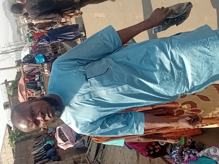A bearded man wearing a blue, patterned long-sleeved shirt stands in an outdoor setting. Behind him, a crowded market scene unfolds with numerous stalls and people milling about. Bags are displayed prominently. In the foreground, a patterned mat lies on the ground near seated individuals. The man is holding a cloth item in his hand.