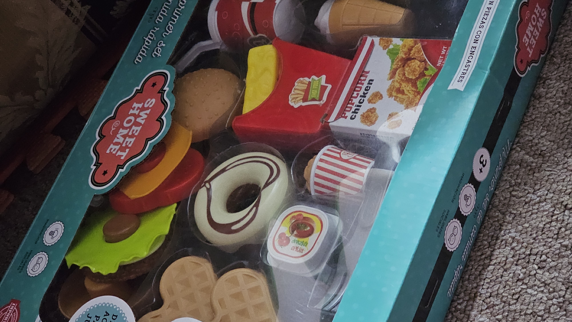 The image shows a toy set of fast food items in a teal box with the words "SWEET HOME" and "Mi primer set de comida rapida" written on it. The set includes a burger, fries, popcorn chicken, a donut, soda, a condiment container, an ice cream cone, and waffles. The box also has symbols for imagination and social skills, with a "+3" symbol indicating the recommended age. The box is placed on a carpeted surface. There are no people in the image, and there is no information related to the location of San Bernardino Tlaxcalancingo, Mexico.