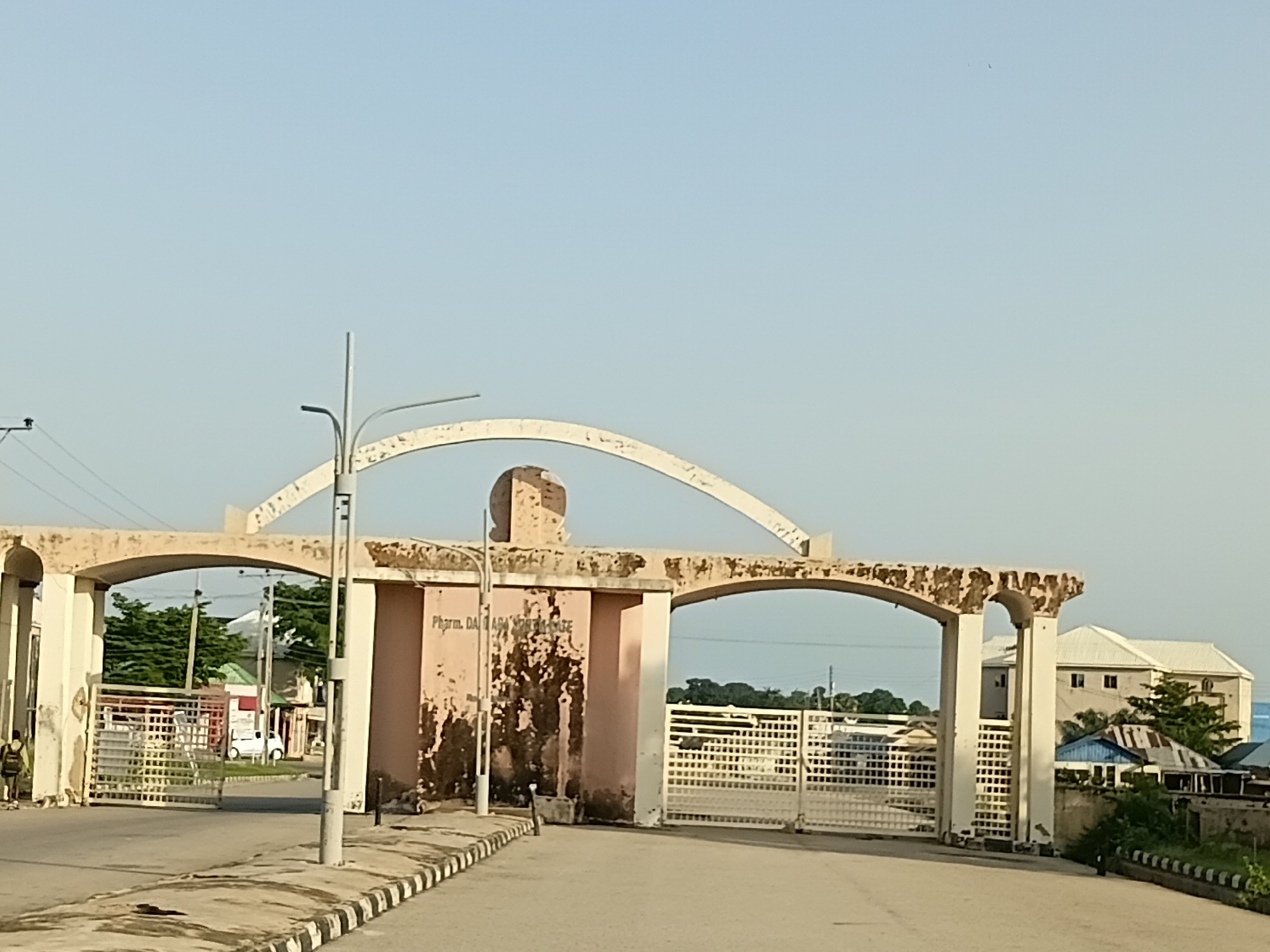 The image depicts the entrance gate to a facility in Jalingo, Nigeria. The gate is made of light-colored, weathered concrete arches and features a white metal grate. The central pillar of the gate has the faded inscription "Pharm. DA DADAY NORTH-EAST". To the left of the gate, a person in dark clothing stands near the entrance. On the road leading up to the gate, there is a white vehicle. Behind the gate, various buildings, including a light-colored multi-story structure with a white roof, are visible amidst greenery. The sky is clear and pale blue. The overall scene is of an institutional entrance under daylight conditions.