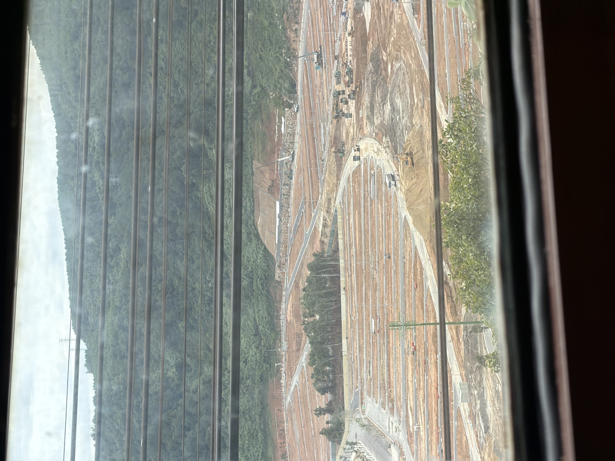The image depicts a large-scale construction site in Hải Vân Ward, Vietnam. The terrain is mountainous and covered in lush green vegetation in the background. In the foreground, the land has been cleared and flattened, with numerous rows of demarcated plots, suggesting the development of a housing or commercial area. Several construction vehicles, including trucks, excavators, and cranes, are actively working on the site. Roads and pathways are being laid out throughout the development. A communication tower is visible in the mid-ground. The scene is viewed through horizontal blinds, which create a striped effect across the image.