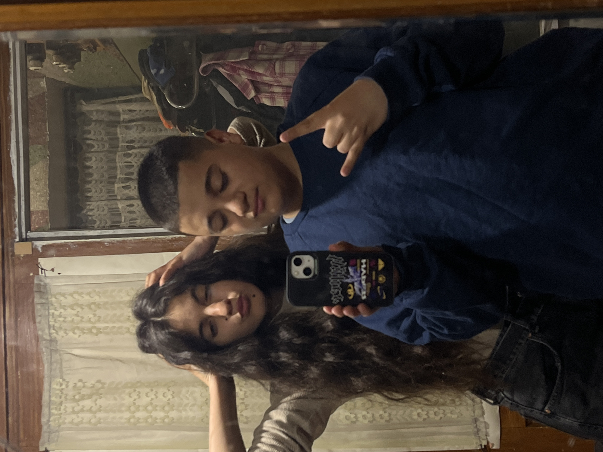 A young man and a young woman are captured in a mirror selfie. The man is positioned at the top of the frame, wearing a blue long-sleeved shirt and making a "rock on" hand gesture. The woman is positioned below him, her long, dark, curly hair cascading around her face. She is holding a smartphone with a distinctive patterned case. The environment suggests an indoor setting, with curtains visible in the background. The location is not visually discernible from the image. There is no way to confirm if the image was captured in Lozarevo, Bulgaria.