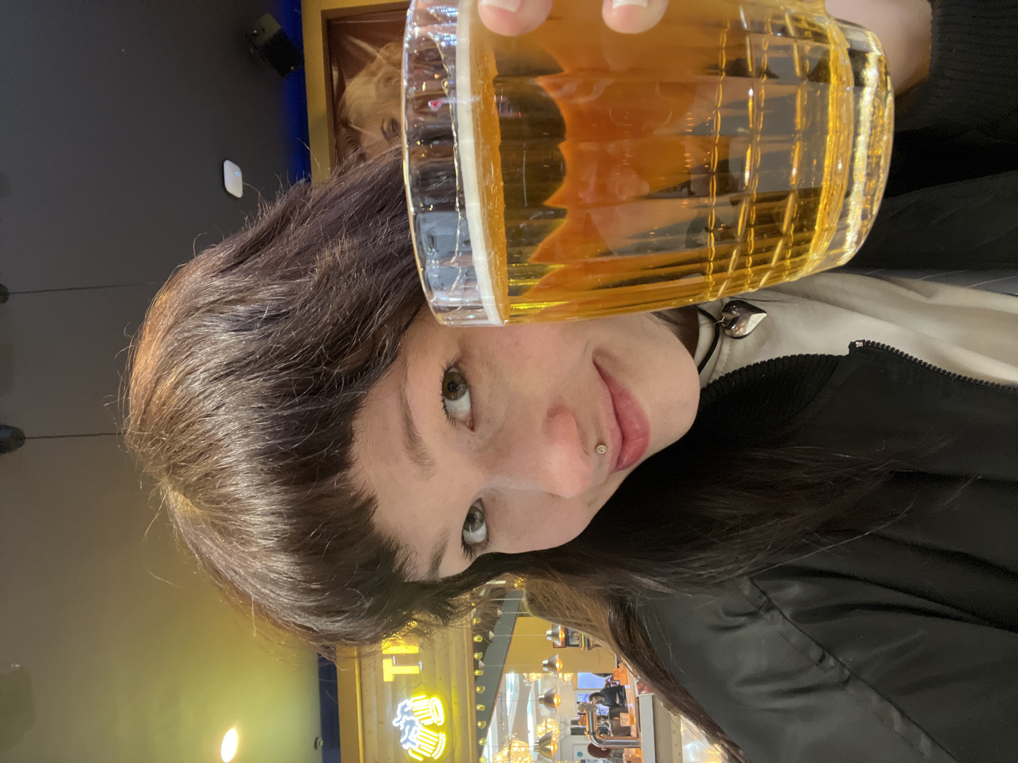 A woman with brown hair and a lip piercing is holding a beer in a glass. She is wearing a black jacket over a white shirt. The background shows a restaurant interior, identifiable as Kazan, Russia by the "TAtNEFT ARENA" logo in neon lights. Other details include pendant lights and tables with people seated.