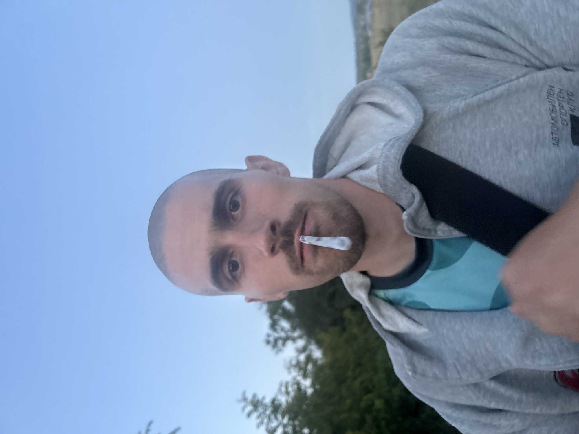 A man with a shaved head and a goatee is shown in a close-up, outdoor, waist-up portrait. He is holding a lit cigarette with a patterned filter between his lips. He wears a grey hooded sweatshirt over a teal and black patterned t-shirt. A black strap, possibly from a bag, is visible across his chest. In the background, blurred green foliage and a distant, muted landscape suggest a park or rural setting. Text is visible on the lower right side of the sweatshirt, appearing to be in Cyrillic, potentially indicating a club or organization. The overall lighting and sky suggest the image was taken during late afternoon or early evening. The text on the sweatshirt contains the words "АВТОМОБИЛЕН СПОРТЕН КЛУБ", which translates to "Automobile Sports Club." While the image is taken outdoors, there is no specific identifying landmark within the frame that would pinpoint a location within Burgas, Bulgaria.