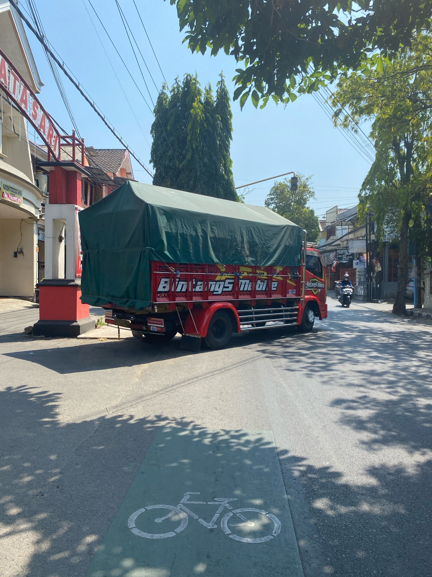 The image shows a red truck with a green tarp covering its cargo, driving on a street in Kediri City, Indonesia. The truck has "BINTANGS MOBIL" written on its side. A motorcycle with a rider wearing a blue helmet is visible in the background, also on the road. The street has a designated bicycle lane marked by a green stripe and a white bicycle symbol. Trees with dappled shadows are present on the right side of the road and overhanging from above, suggesting it is a sunny day. Buildings are visible on the left side of the street, with one building displaying signage that appears to be in Indonesian. Overhead wires crisscross the sky.