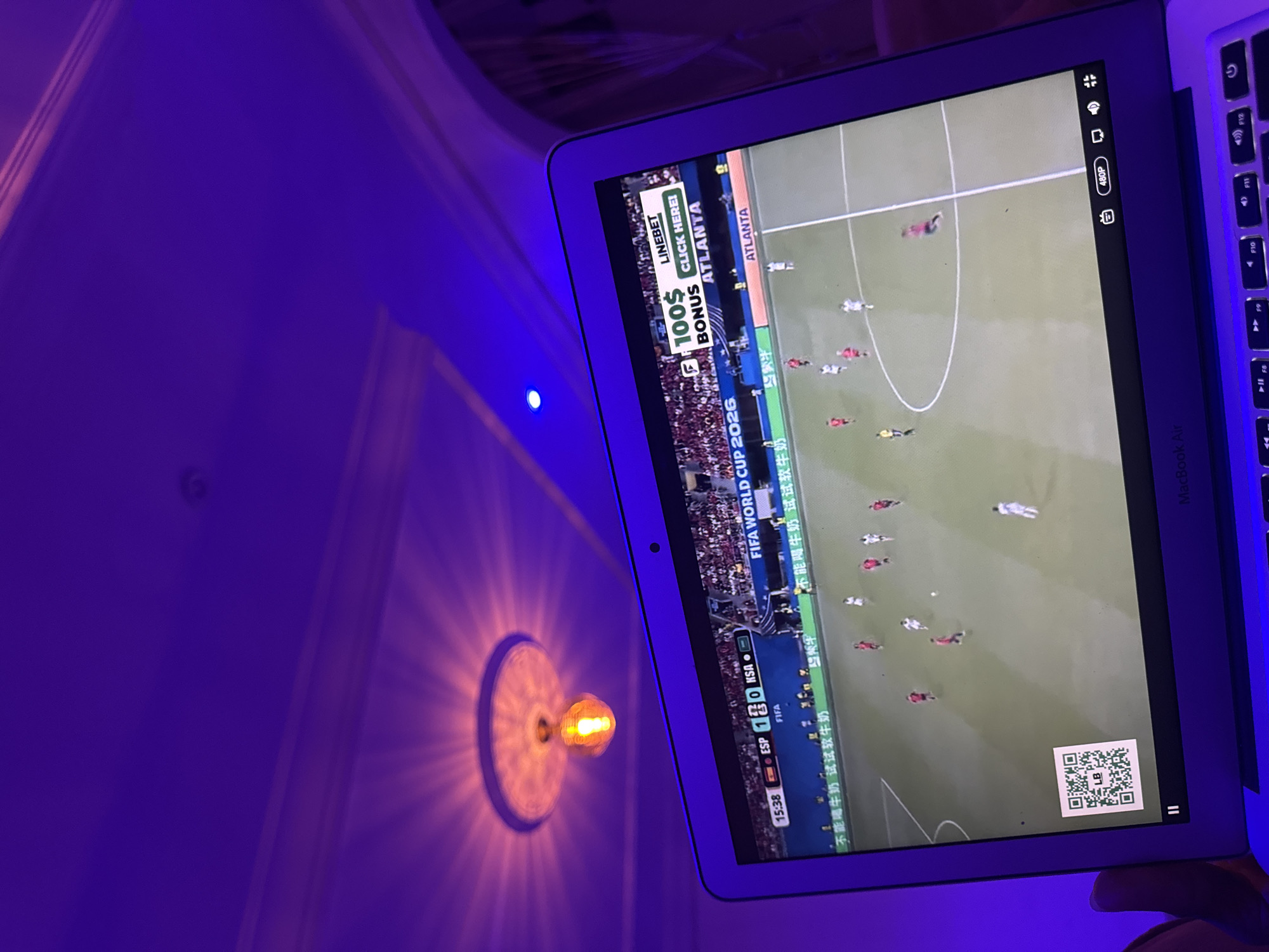 The image shows a close-up of a laptop screen displaying a soccer match. The screen is illuminated by purple ambient lighting, casting a glow on the laptop's keyboard and the surrounding ceiling. On the screen, a soccer game is in progress, with players in red and white uniforms on a green field. A scoreboard at the top of the screen indicates "ESP 1 0 KSA", suggesting a match between Spain and Saudi Arabia. Above the game, an advertisement for "LINEBET" with "$100 BONUS" and "CLICK HERE!" is visible, along with the text "ATLANTA". In the bottom left corner of the screen, a QR code is displayed. The laptop appears to be a MacBook Air, as indicated by the branding on the keyboard. The overall scene suggests a person is watching a soccer match on their laptop in a room with purple mood lighting, possibly in Laminga, Nigeria.