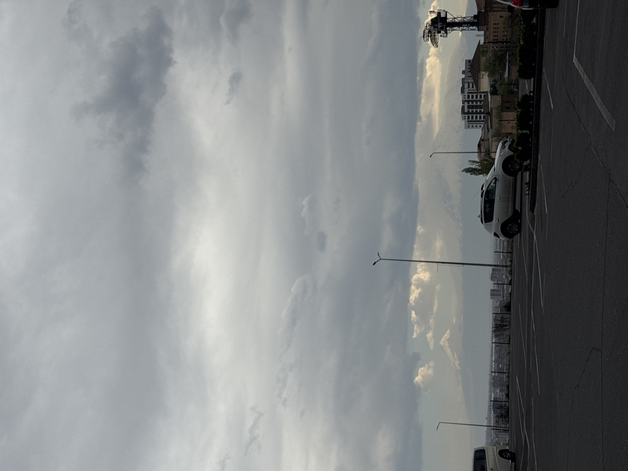 The image shows a view from a parking lot in Yerevan, Armenia. The right side of the image shows a parking lot with black asphalt and white parking lines, with several parked cars including a white SUV. Behind the parking lot is a view of the city with buildings of varying heights. In the upper left corner, there is a sky filled with gray clouds, suggesting an overcast day. There is also a tall metal tower visible on the left side of the image. Streetlights stand intermittently throughout the scene.