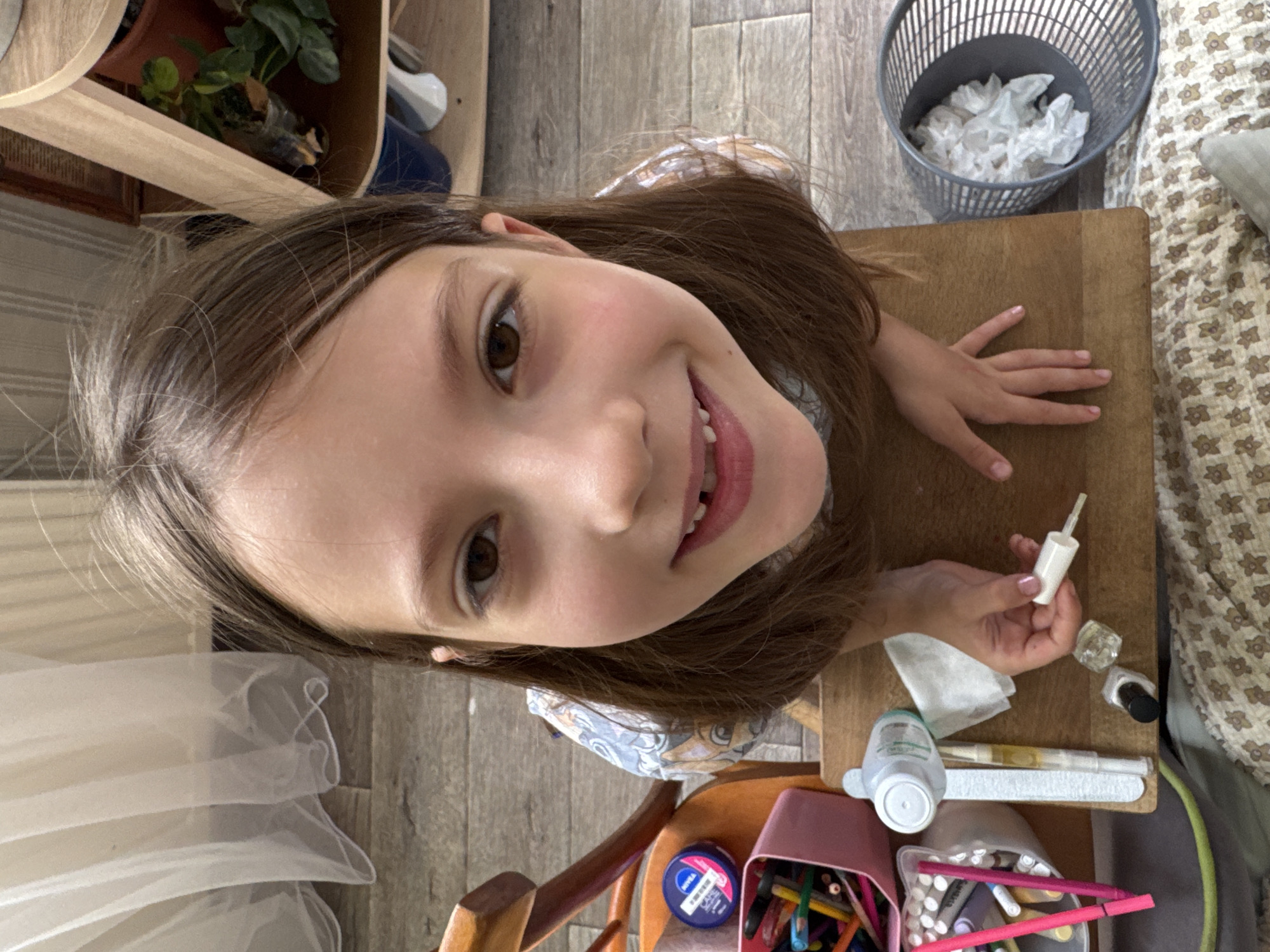 This is a candid, overhead shot of a young girl sitting at a wooden table, engaged in what appears to be a nail art activity. The girl is the primary subject, with her face filling a significant portion of the frame, looking directly at the camera with a bright, open-mouthed smile and a mischievous glint in her brown eyes. Her long, brown hair frames her face, with some strands falling forward.
On the table in front of her are various items associated with nail care and art. These include a white bottle with a brush applicator, a small glass bottle, a dark-colored nail polish bottle, a white tissue, a bottle with a green cap, and a nail file. There is also a pink container filled with colorful markers or colored pencils. A small blue jar with white and pink text is visible, which appears to be a cosmetic product, possibly Nivea.
The background shows a wooden floor and a portion of a patterned fabric, possibly a blanket or bedding. To the upper right, a grey mesh trash can is filled with crumpled white tissues. In the upper left corner, a shelf with a potted plant is visible. The lighting suggests it is daytime. The overall mood is cheerful and playful, captured during a moment of personal grooming and creative expression. The visible text on the blue jar is "NIVEA" and "CREME". The location is Zassovskaya, Russia.