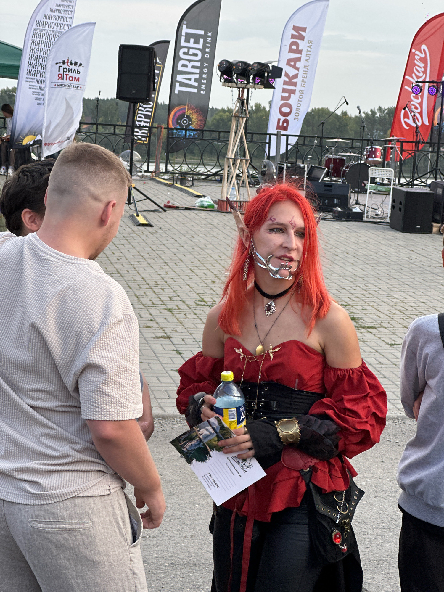 A young person with bright red hair, elf-like ears, and facial piercings is standing on a paved area, possibly a city embankment, holding a water bottle and a pamphlet. They are wearing a red off-the-shoulder top with puffy sleeves, a black corset, and black pants. In the background, there are several flags with Russian text and logos, a stage with musical instruments and lighting equipment, and a body of water. The weather appears to be overcast. The scene suggests an outdoor event or festival.