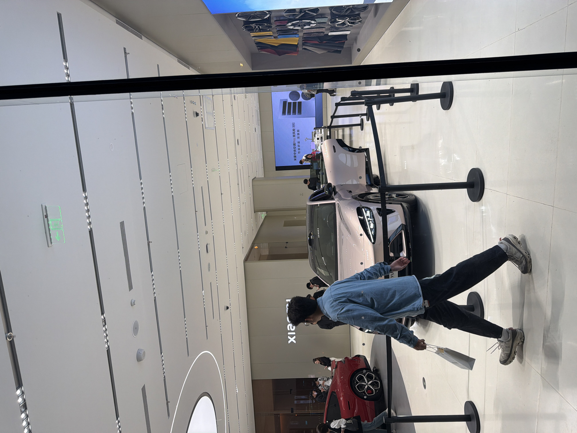 The image shows a person wearing a light blue denim jacket, black pants, and gray sneakers walking in what appears to be an automotive showroom in the Jiangning District, China. Several cars are on display, including a pink car with a door ajar and a red car. The person holds a light-colored bag. Barriers with black poles and bases are set up around the cars. A large display screen is in the background, along with the word "Xiaomi" on a wall. The showroom has a modern design with a bright, white ceiling featuring recessed lighting. The image is taken through a window or glass panel.