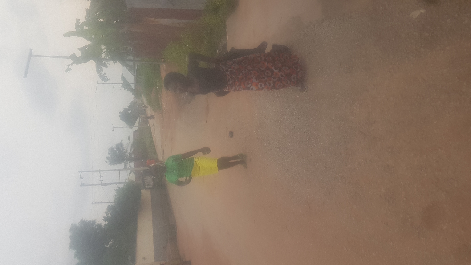 The image depicts an outdoor scene on a dirt road in Wukari, Nigeria. Two young women are the central figures. The woman on the left wears a green t-shirt and a yellow skirt, and she is looking towards the right. The woman on the right is wearing a dark top and a patterned skirt, and she is facing away from the camera, with her right arm raised. There are other individuals and a vehicle faintly visible in the background. The road is unpaved and appears to be surrounded by vegetation and buildings. The sky is overcast, suggesting cloudy weather.