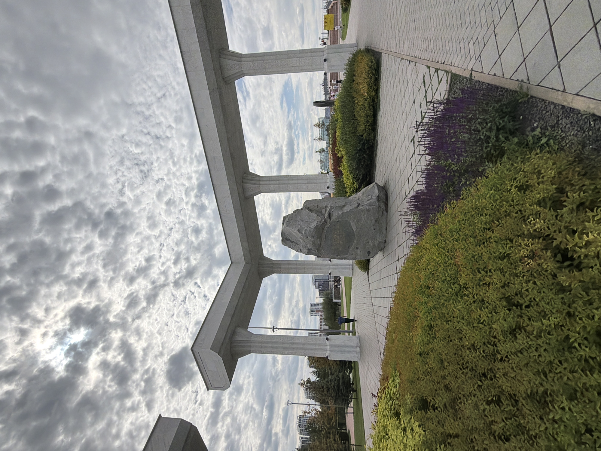This image depicts a public space in Astana, Kazakhstan, likely a park or plaza. The foreground is dominated by dense, manicured green shrubs and a cluster of purple flowering plants, bordering a paved walkway composed of light gray, square tiles laid in a diagonal pattern. To the right of the walkway, more of the same tiled paving extends into the distance.
In the mid-ground, a large, rough-hewn grey boulder sits prominently on a circular tiled area, appearing to be a monument or art installation. Behind the boulder and walkway, a modern architectural structure with large, fluted white columns and a white horizontal beam stretches across the scene, creating a covered walkway or archway. A single person in a dark shirt and light pants is walking away from the camera down the tiled path under this structure.
The background reveals a cityscape with numerous modern high-rise buildings, some of which have a white facade. Trees with green and autumnal foliage are visible between the buildings and the foreground structures. The sky is dramatic, filled with a dense layer of textured, gray and white clouds, with a bright spot indicating the sun partially breaking through. The overall impression is one of a contemporary urban environment with a focus on landscaping and monumental elements.