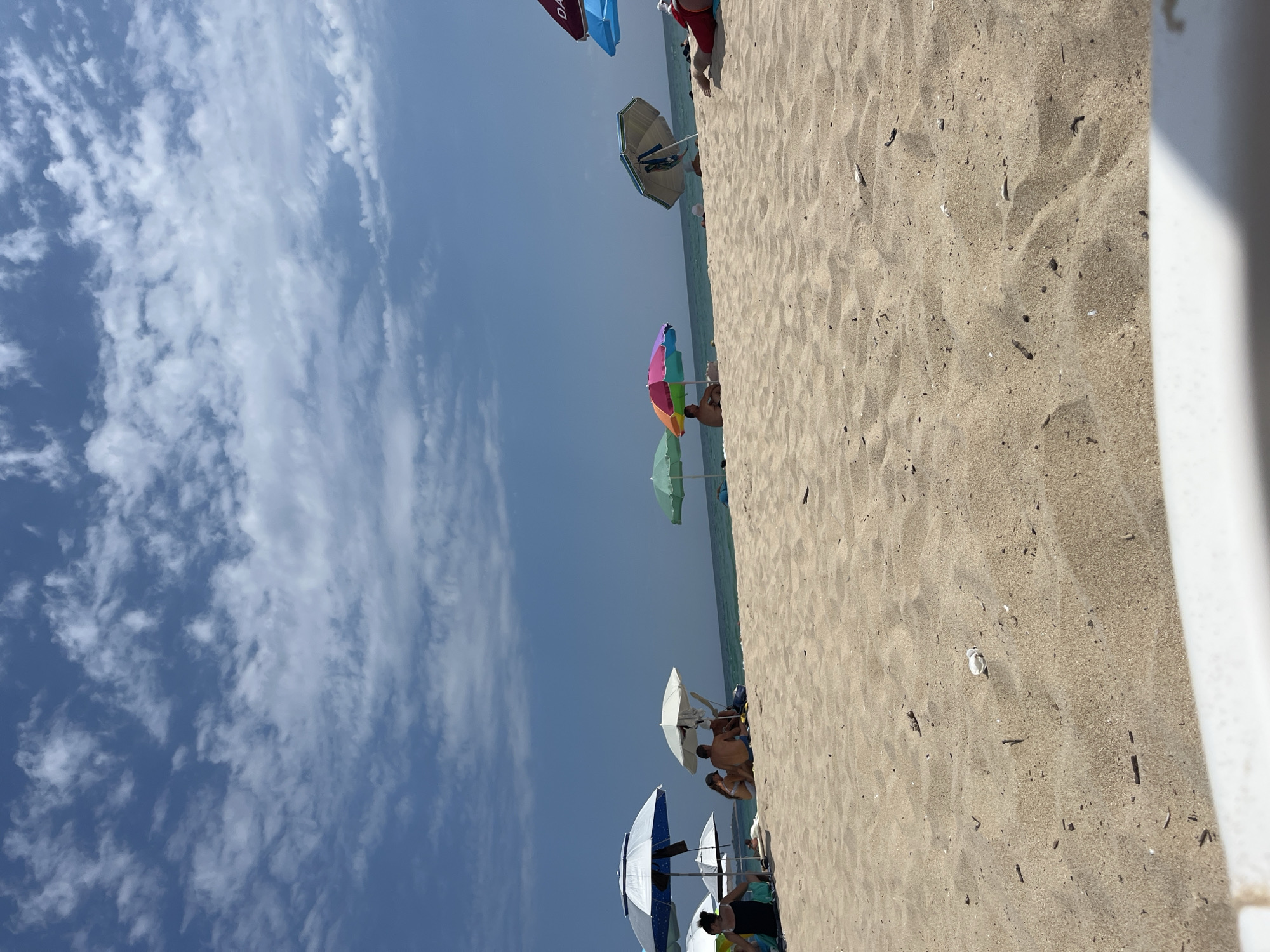 A group of people are gathered on a sandy beach under a partly cloudy sky. Several beach umbrellas are set up on the sand, providing shade. Some individuals are sitting or lying down. The sea is visible in the background, meeting the horizon under the sky. The scene appears to be a typical summer day at a coastal location.