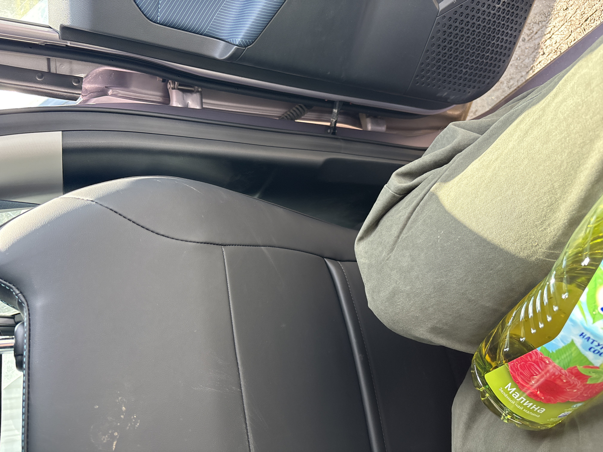 The image captures a partial view of a car's interior on a sunny day, likely in Izhevsk, Russia. The main subjects are a person's leg, a car seat, and a beverage bottle.
A person, whose lower half is visible wearing olive green pants, is seated in the car. Their leg is positioned to the right, holding a clear plastic bottle. The bottle contains a yellowish-green liquid and has a label featuring images of raspberries and green leaves. Visible Cyrillic text on the label reads "Малина" (Raspberry) and "Зеленый чай малина" (Green tea raspberry). Partially visible text includes "НАТУРАЛЬНЫЙ СОК" (Natural juice).
The car interior features a dark, possibly leather or synthetic leather, seat with visible stitching and some light marks or stains on its surface. The car door is open, revealing part of a speaker grille with a hexagonal pattern and a section of the door panel with a light blue patterned fabric. Bright sunlight streams into the car, indicating daytime, and highlights the textures of the fabric and plastics. The ground outside the open car door is also visible, suggesting the car is stationary outdoors.