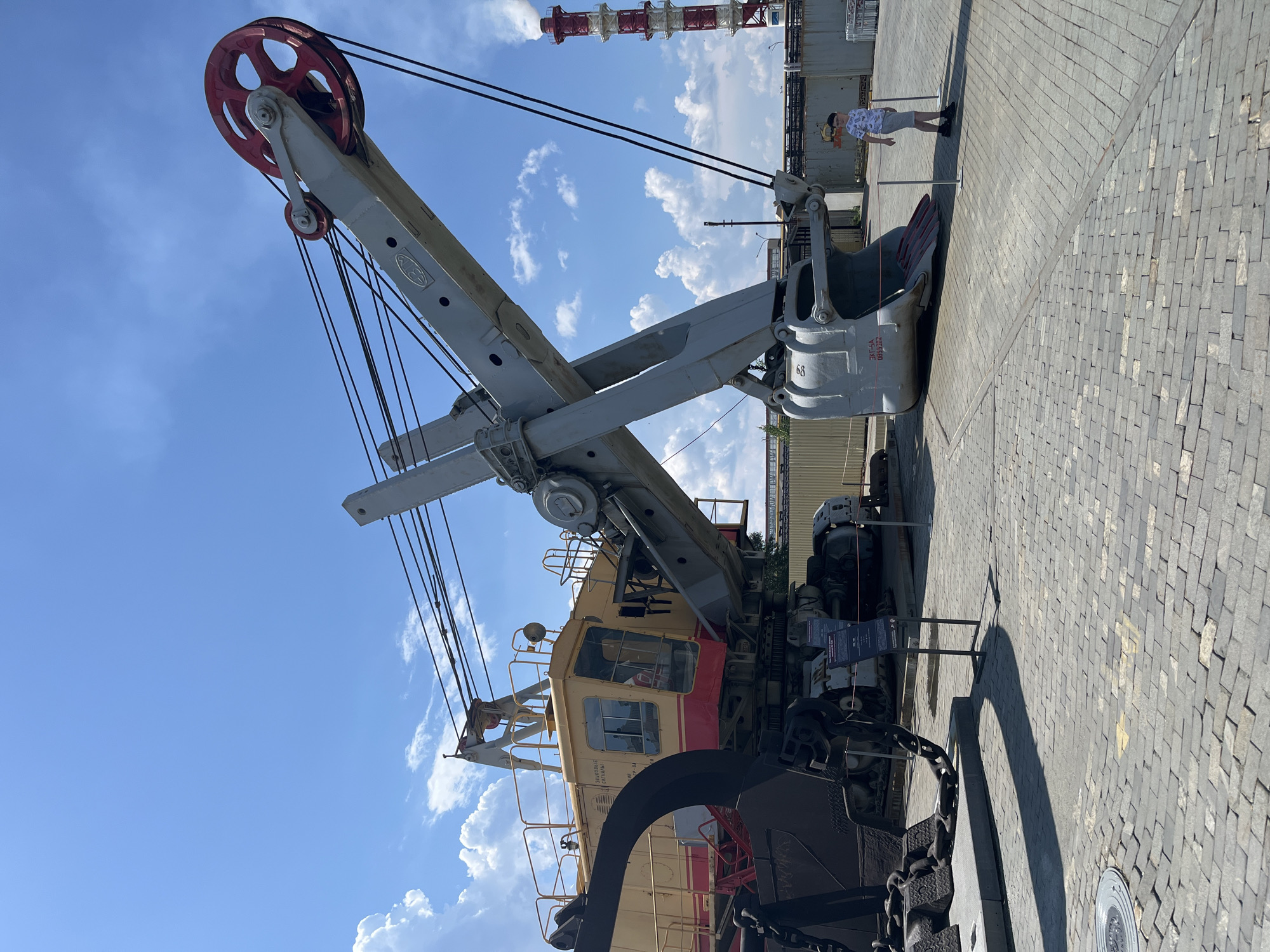 In Verkhnyaya Pyshma, Russia, at the Museum of Military Equipment "Battle Glory of the Urals," a massive, yellow and red excavator is displayed on a paved area under a blue sky with scattered white clouds. The excavator, a large industrial piece of machinery, features a prominent boom with a large red pulley at the top and a substantial grey bucket hanging from its end. The machine is complex, with numerous cables and structural elements visible. To the right of the excavator, a young boy wearing a patterned t-shirt and shorts stands near a red and white striped industrial chimney emitting a plume of white smoke, suggesting the presence of a power plant or factory in the background. The ground is paved with grey cobblestones. A small informational sign is positioned to the left of the excavator's bucket. The scene appears to be an outdoor exhibition, with a sense of scale conveyed by the immense size of the machinery compared to the human figure.