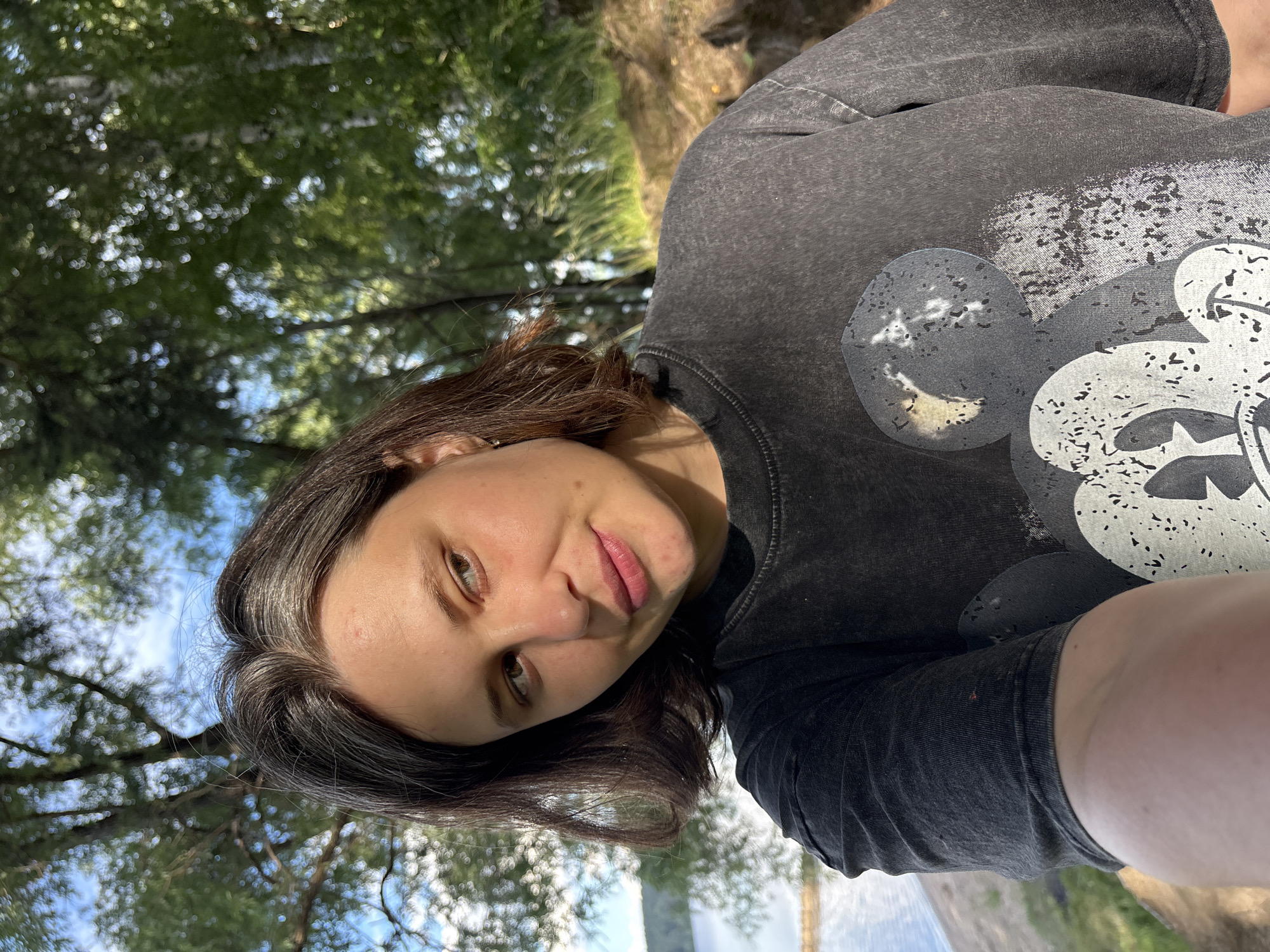 The media file shows a selfie of a young woman. She has dark brown hair and is wearing a dark gray t-shirt with a graphic of Mickey Mouse on it. The background is out of focus, but appears to be a natural setting with trees and sunlight. The woman is looking at the camera with a slight smile. The image is taken outdoors, and the lighting suggests it is daytime. The setting appears to be a park or wooded area, possibly near a body of water given the hint of blue in the background. The woman's expression is relaxed and content. There is no visible text in the image.
