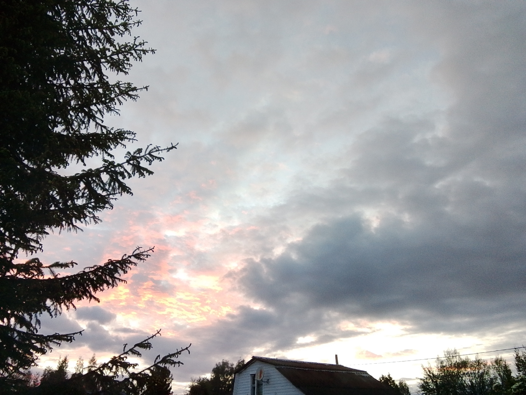 The image shows a cloudy sky with a hint of pink and orange hues, possibly at sunset or sunrise. A large pine tree is present on the left side of the image. Below the clouds and to the right, a small building with a dark roof and what appears to be a satellite dish on the side is visible. The location in the city of 1-Go Maya, Russia is not verifiable from the image.