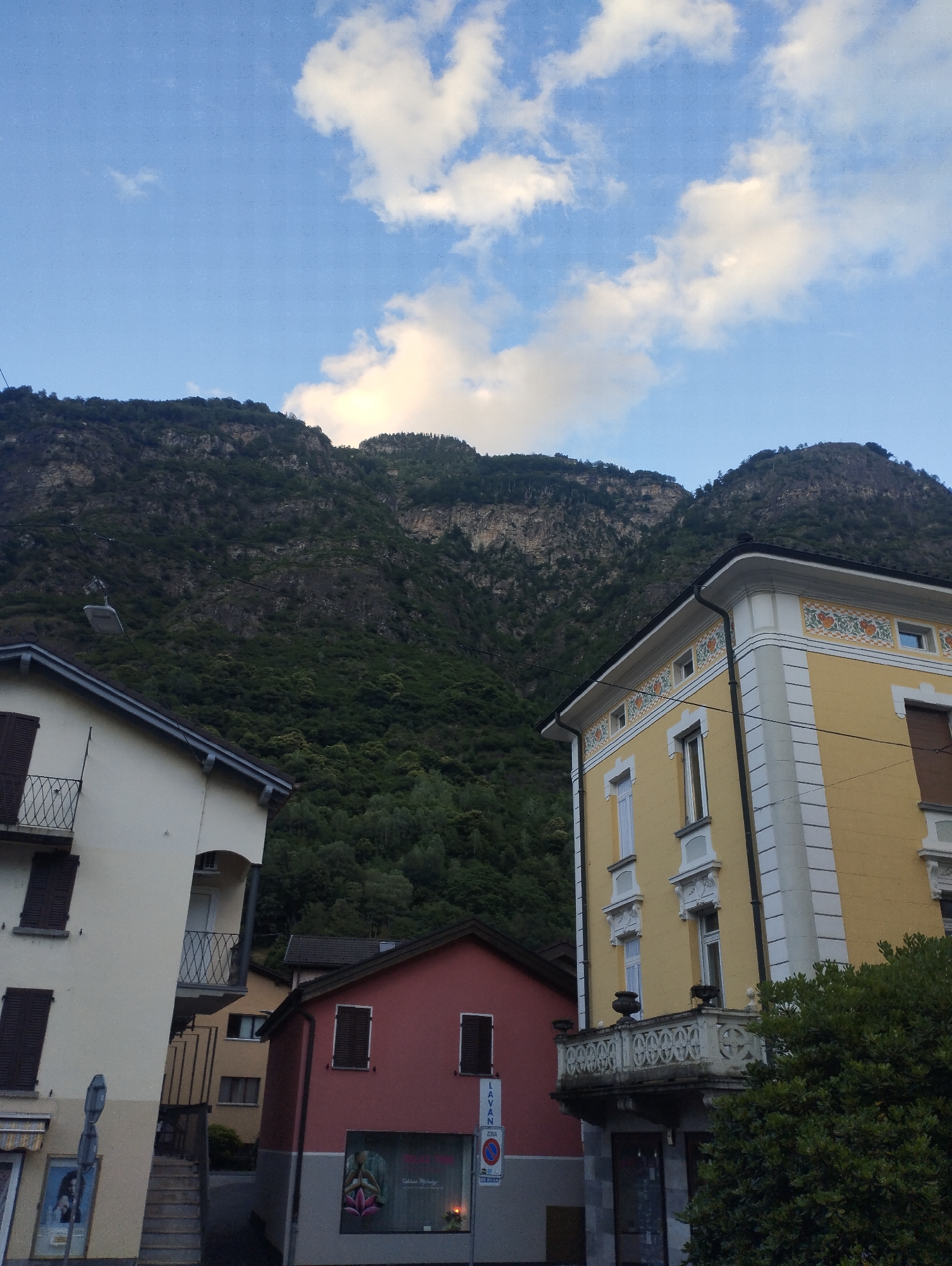 The image shows a view of buildings in Biasca, Switzerland, with a steep, tree-covered mountain in the background. In the foreground, there is a cream-colored building with dark shutters on the left, and a yellow building with decorative white trim and ornate balconies on the right. A smaller red building is situated between them. The sky above is blue with scattered white clouds. A street lamp is visible hanging from a wire in front of the mountain. The scene appears to be in an urban or semi-urban area, possibly a town or village nestled at the base of the mountain. There are no people visible in the image. The overall impression is one of a picturesque landscape where natural scenery meets architectural structures.