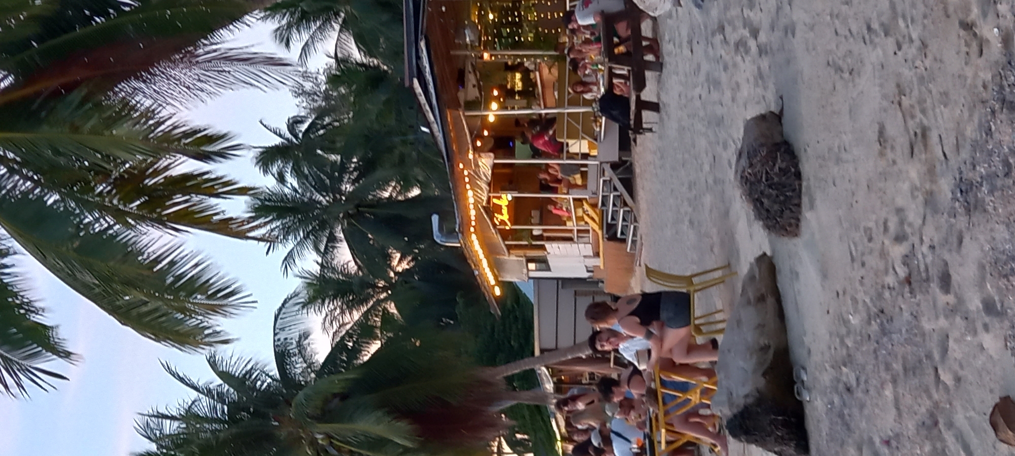 The image depicts an evening scene at a lively, open-air establishment, likely a restaurant or bar, situated in a tropical location, specifically Baan Sri Thanu, Thailand. The setting appears to be beachside or very close to the coast, with light-colored sand or dirt forming the ground in the foreground and surrounding the structure.
Dominant features include numerous palm trees, their fronds visible against a pale sky, suggesting the transition from dusk to early evening. The multi-level building is well-lit with numerous string lights, creating a warm and inviting ambiance. A notable detail on the building's facade is a neon sign, possibly spelling "Thank you" or "Tunku" in a cursive script.
Numerous people are present, socializing and relaxing. They are seen on both levels of the building, seated at tables, and standing in groups. Many appear to be enjoying drinks and conversations in a casual atmosphere. Their attire is informal, suited for a warm, outdoor environment. A yellow plastic chair is visible on the sandy ground near the building. The overall impression is one of a relaxed gathering, enjoying the early evening hours in a tropical resort or beach area.