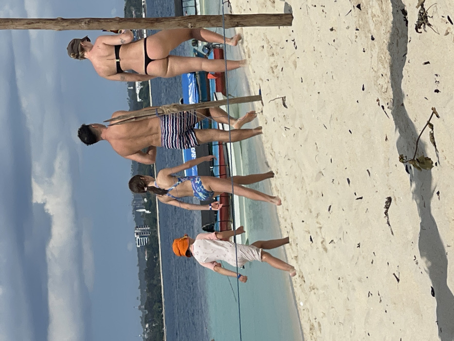 A sandy beach scene in Zanzibar City, Tanzania, shows four individuals walking barefoot from left to right along the shoreline under a cloudy sky. The foreground consists of light-colored sand with scattered dark debris. From left to right: a young child in an orange bucket hat and a light-colored rashguard; a young girl with dark hair in a ponytail wearing a blue patterned one-piece swimsuit; a bare-chested man in blue and white striped swim trunks carrying a long wooden pole over his right shoulder; and a woman in a black bikini top and thong bottom holding a brown bottle in her right hand. A blue rope stretches horizontally across the mid-ground, and two thick, light-colored wooden poles stand vertically on the right. Behind the individuals, several small, brightly colored boats (red, blue, white) are partially visible in clear, shallow turquoise water. The water extends into a deeper blue, with a distant coastline visible across the bay, featuring green vegetation and a city skyline with several multi-story buildings.