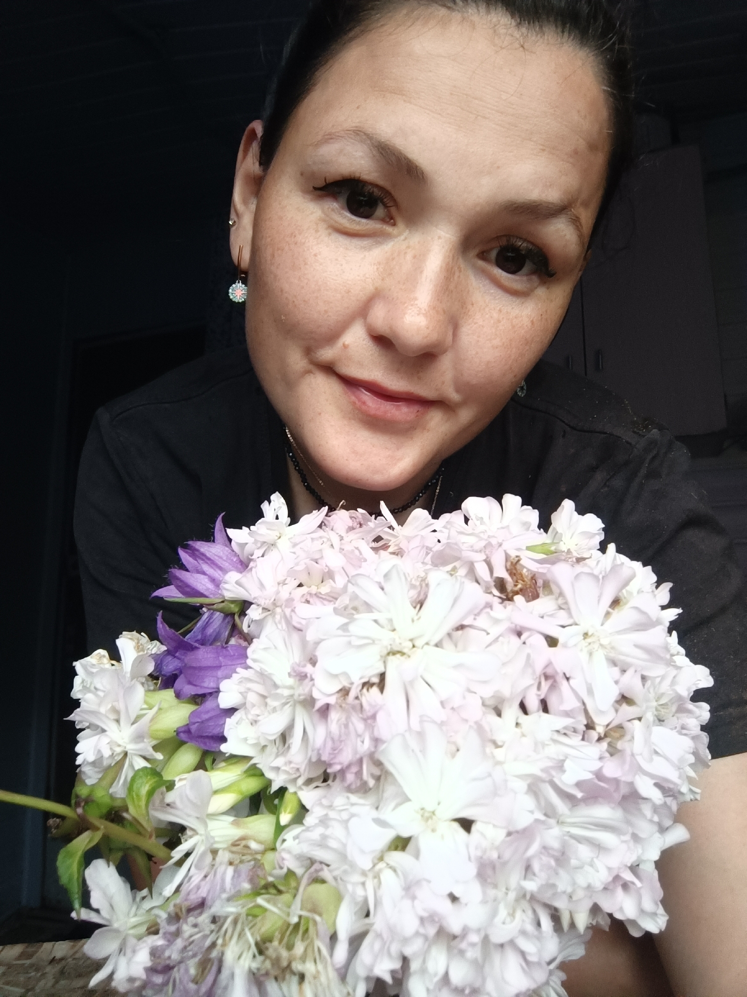 A close-up, head-and-shoulders portrait shows a woman with dark hair pulled back and fair skin with freckles. She is wearing a black t-shirt and a delicate necklace. She is holding a bouquet of light-colored flowers, predominantly white with some purple accents, in front of her. The background is dark and out of focus, suggesting an indoor setting. The image appears to have been taken in Karsha, Russia.