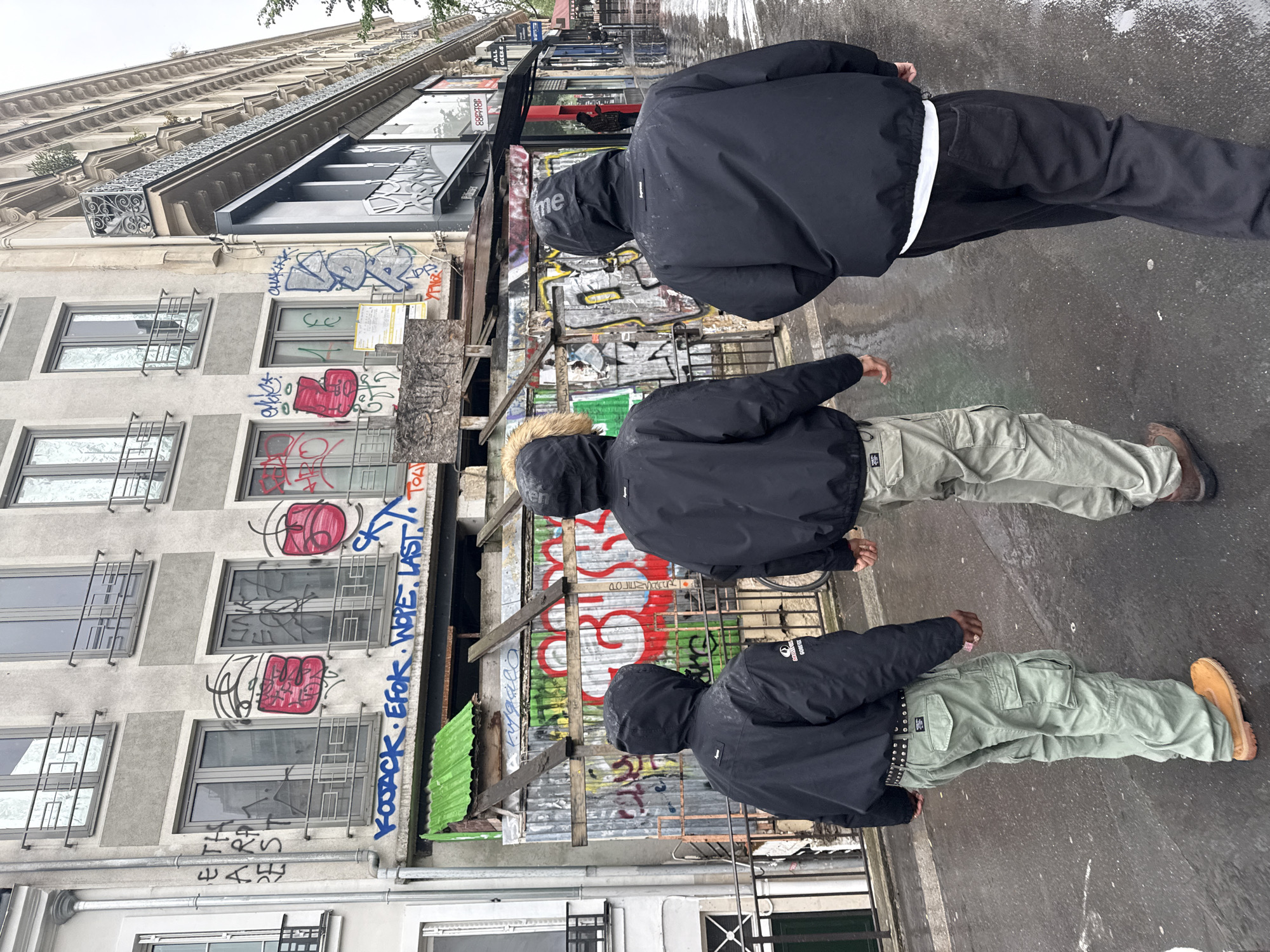 Three figures walk side-by-side on a wet street in Paris, France. The person closest to the camera wears a black jacket and light green cargo pants with a studded belt and tan boots. The middle figure is wearing a black jacket, light green cargo pants, and brown shoes. The person furthest from the camera wears a black jacket and pants with a white stripe. The architecture surrounding them is a mix of old and modern buildings with graffiti art. The street appears wet from a recent rain.