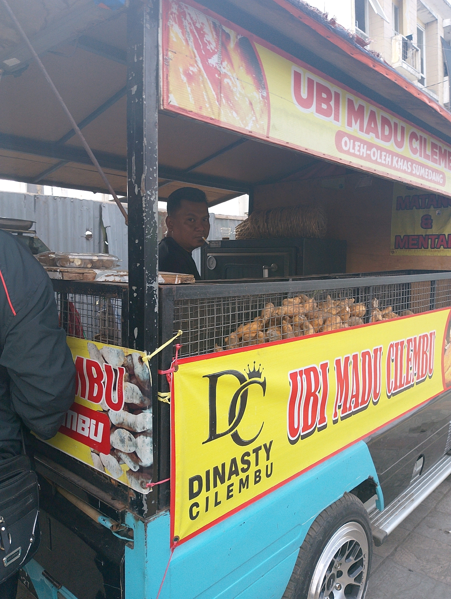 A street food cart in Lembang, Indonesia, is pictured. A man with short dark hair is seen from the chest up, wearing a black shirt, and smoking a cigarette. He is standing behind a metal cage filled with root vegetables. In front of the cart, a large yellow banner displays text in red and black. The text on the banner appears to be a product name, possibly referring to "Ubi Madu Cilembu" (sweet potato of Cilembu). The cart itself is painted in blue and black, and it has chrome trim around the wheels. The background shows a building with balconies and a metal fence.