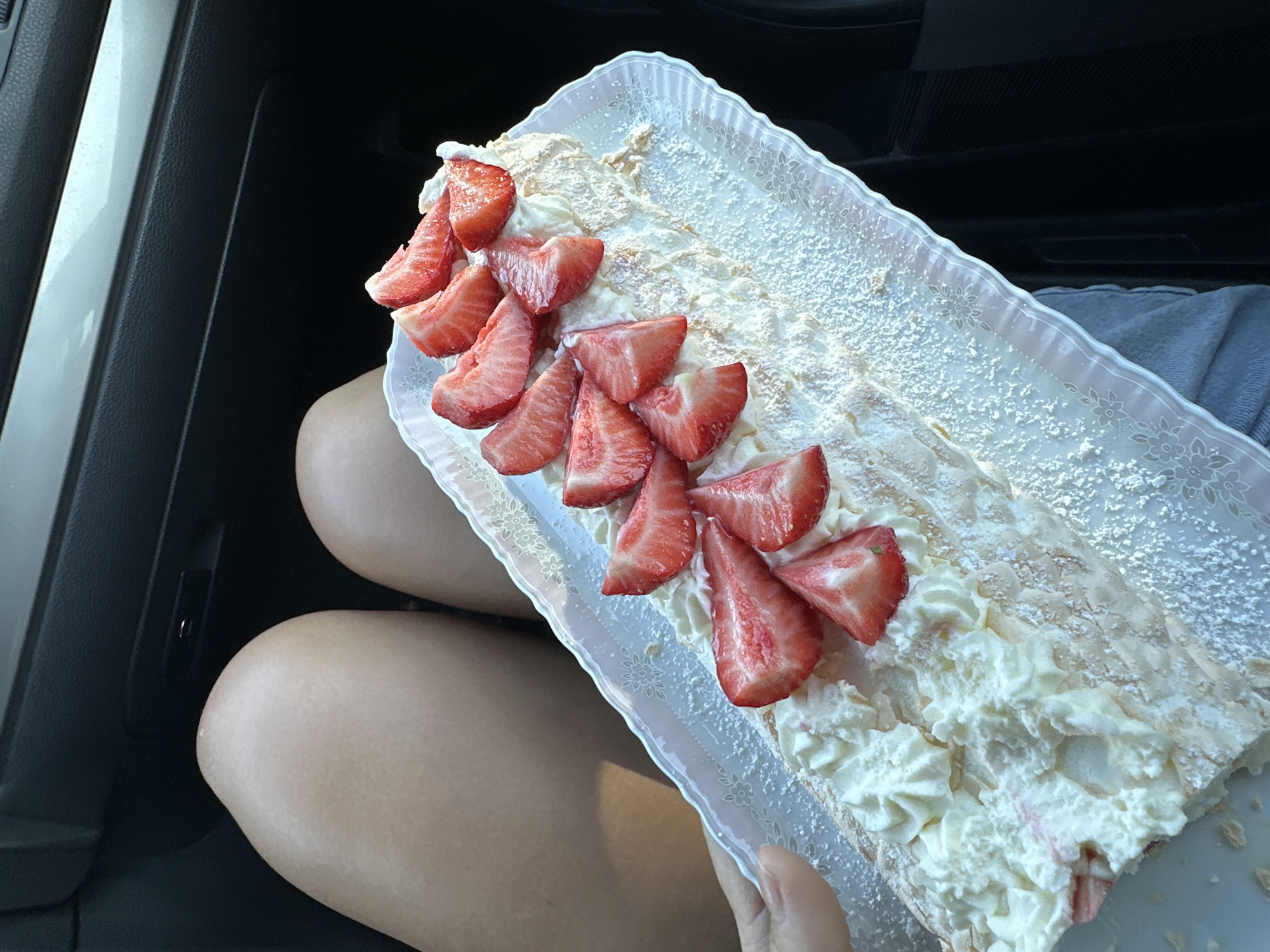 A photograph taken indoors, likely in a vehicle, shows a rectangular, white, decorative plastic tray holding a meringue roll cake decorated with sliced strawberries and dusted with powdered sugar. The tray rests on a person's lap, composed of two bare legs. The background includes a dark car interior. The location is not discernible from the image, so it cannot be confirmed to be in Kutenholz, Germany.