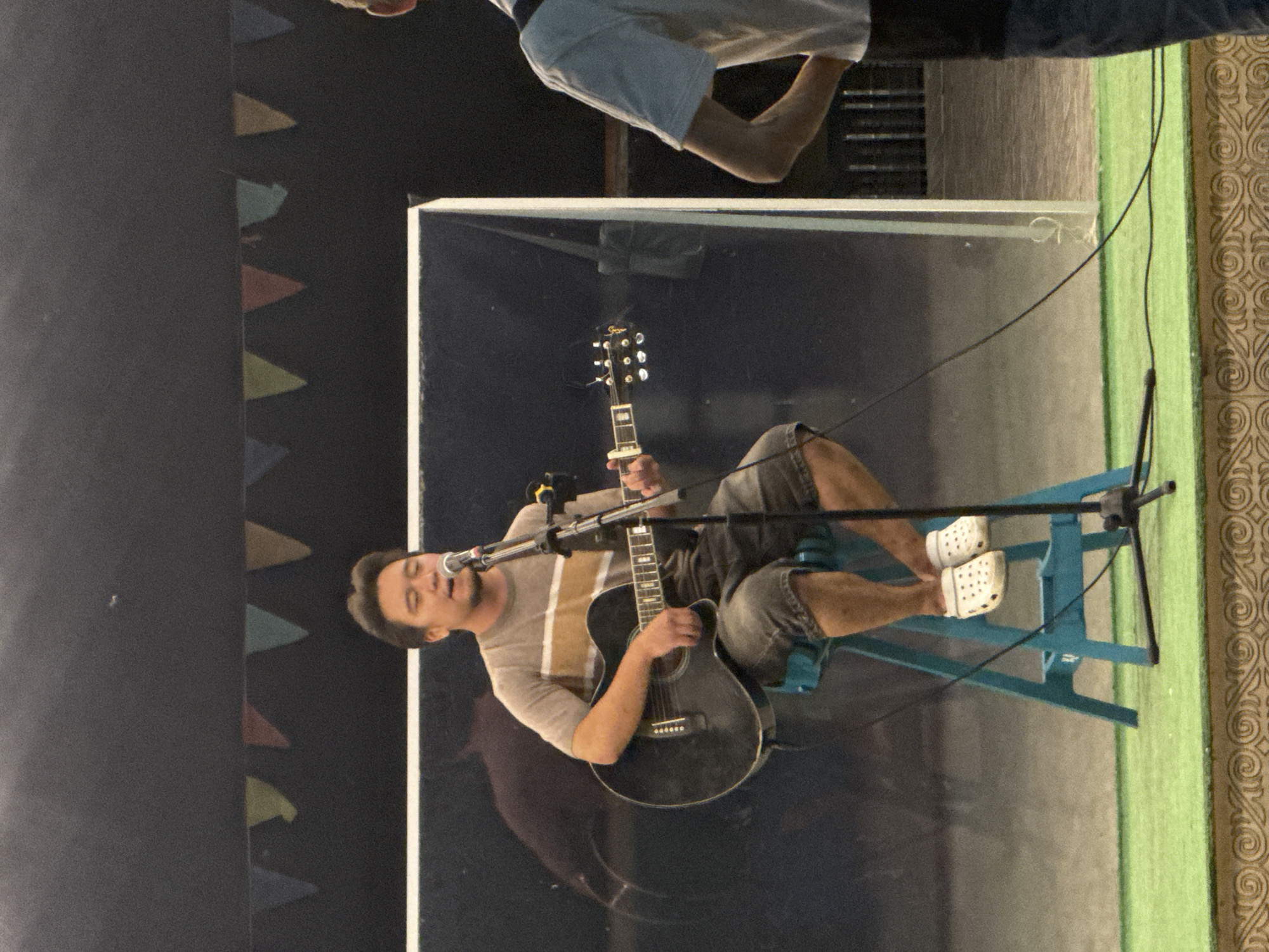 The image shows a man playing an acoustic guitar and singing into a microphone while seated on a blue stool. He is wearing a striped t-shirt, shorts, and white Crocs. In the background, there is a large black panel with a reflection, and colorful triangular flags are strung across the top. To the right, the back of another person wearing a white t-shirt is visible. The foreground shows a green carpeted stage with decorative edging. The scene appears to be an outdoor performance. The location is not specified within the image or its context, and therefore cannot be identified as Pavlodar, Kazakhstan.