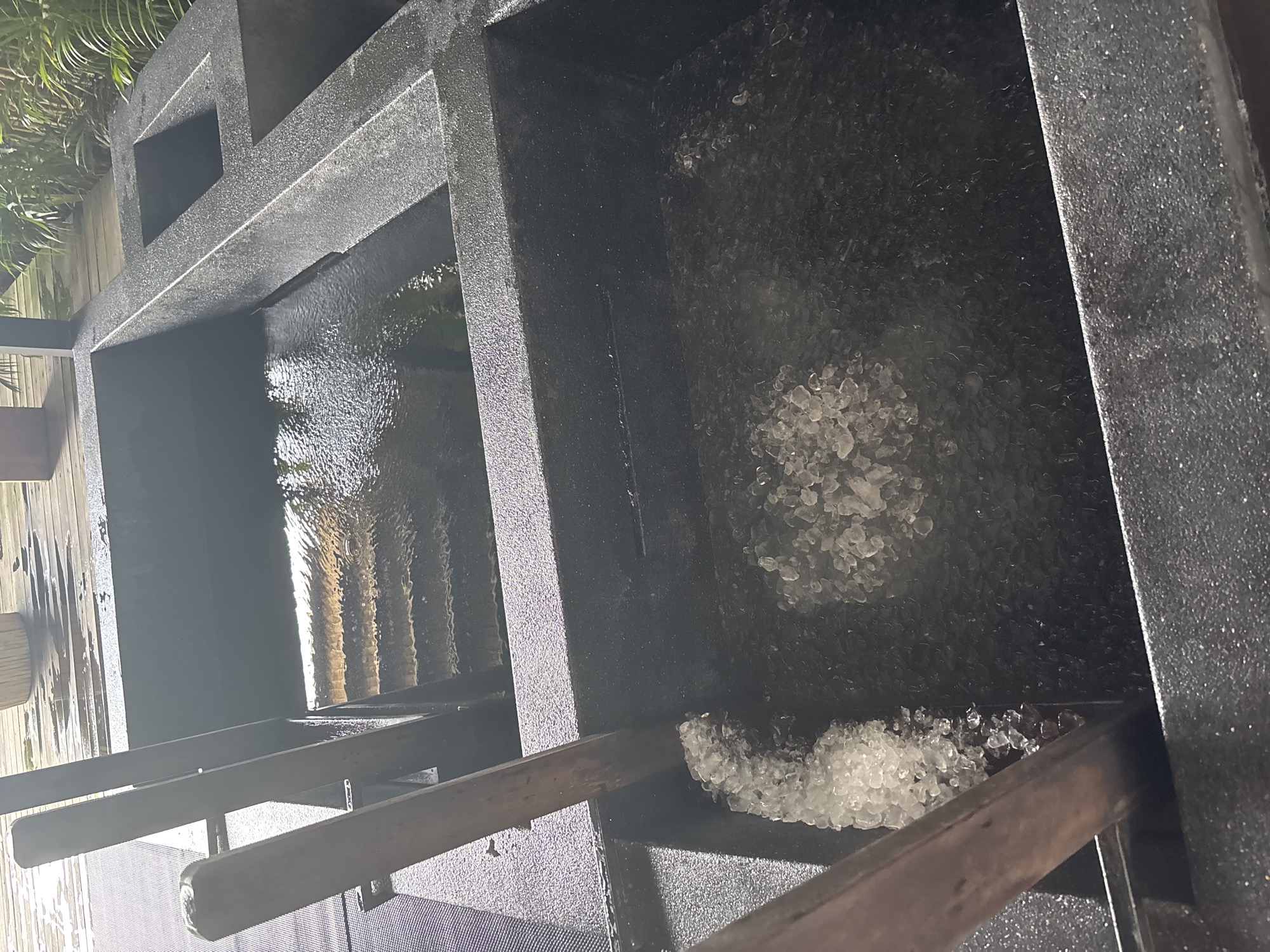 The image shows a close-up view of two large, dark, rectangular basins. The basin in the foreground is filled with a layer of crushed ice. A wooden paddle is partially submerged in the ice, suggesting recent use. The basin in the background contains dark water with ripples, indicating movement. The basins appear to be made of a speckled, rough-textured material. The scene is outdoors, with a wooden deck visible in the background. Lush green foliage can be seen to the right, suggesting a tropical or semi-tropical environment. The lighting suggests it is daytime. The location is identified as Ban Bang Rak, Thailand.