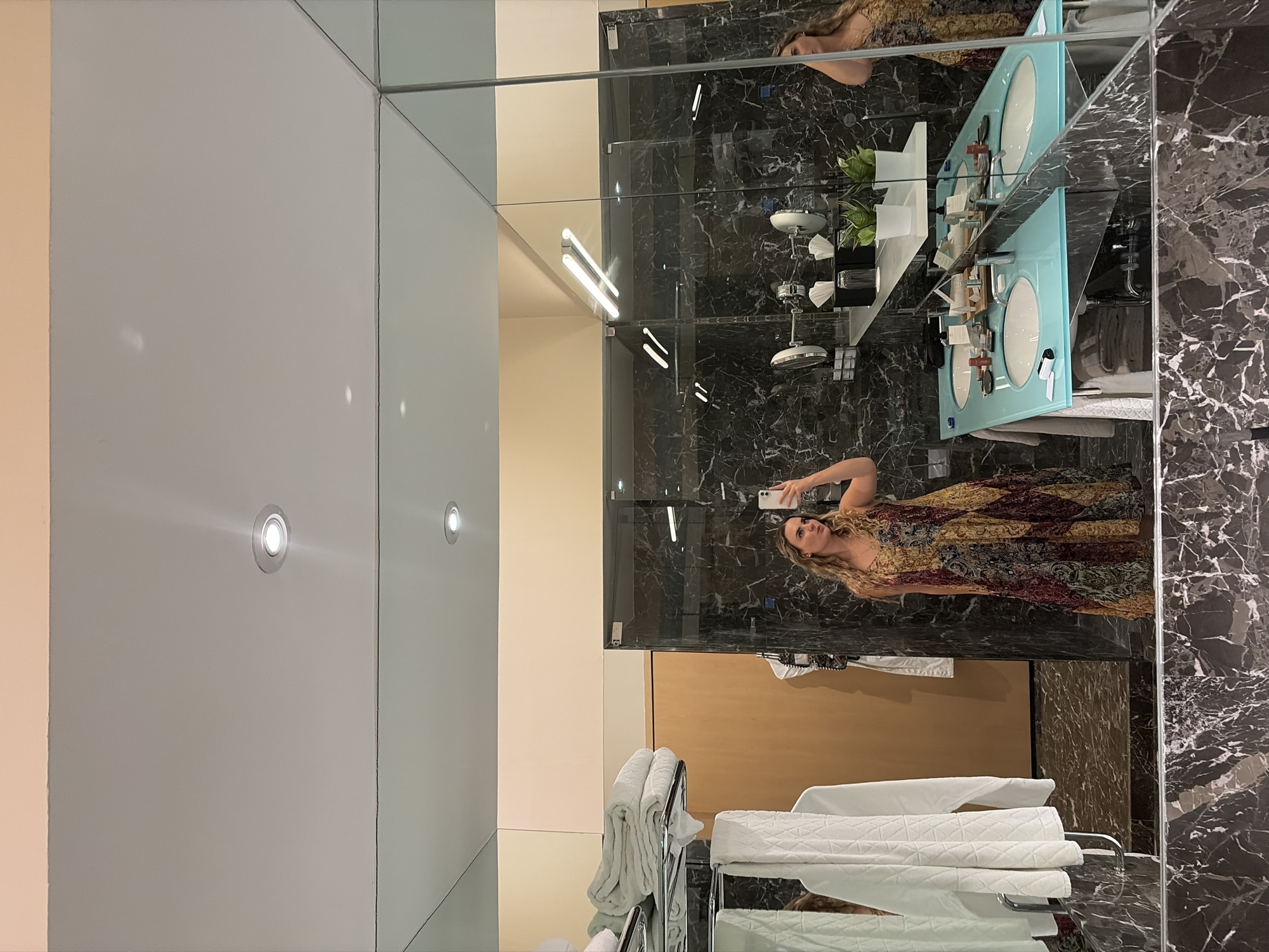 The image shows a reflection in a mirror in a bathroom, possibly in a hotel or upscale residence. A woman is visible in the mirror taking a selfie with her phone. She has long blond hair and is wearing a long, patterned dress in colors of brown, gold, and burgundy.
The bathroom features a marble-patterned wall. A light-blue, double-sink vanity is visible, with various bathroom items on the counter. A metal towel rack holding white towels is in the foreground. The walls are a light neutral color, and there are recessed lighting fixtures in the ceiling.
Given the style and details, the location could potentially be near the Hotel Marqués de Riscal in Elciego, Spain, though this is based solely on the visual characteristics of the bathroom and not explicitly stated within the image.