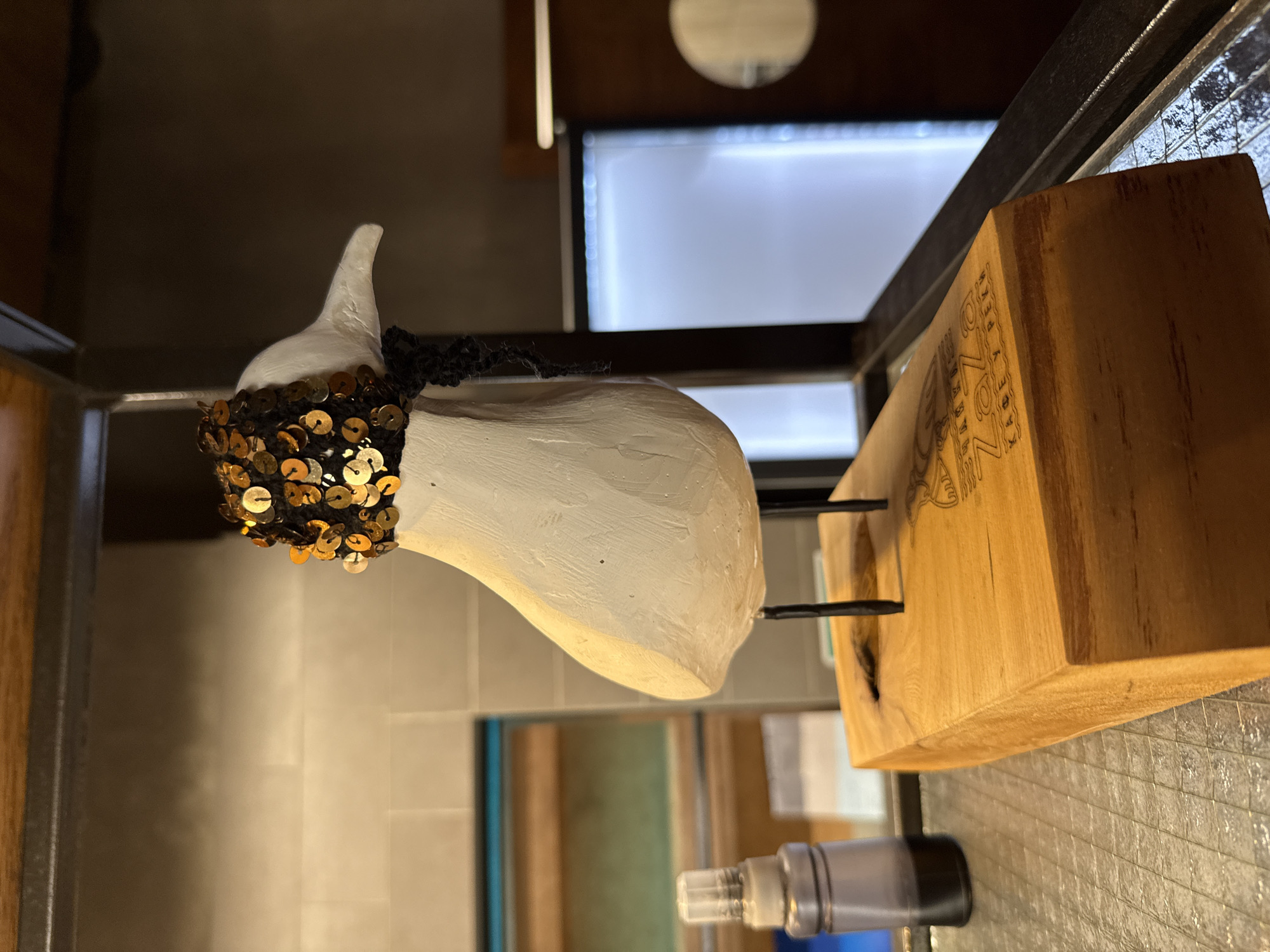 A close-up, eye-level shot captures a decorative, white wooden bird figurine perched on a light brown wooden base. The bird has thin black legs and is adorned with a dark knitted scarf around its neck, topped with a black knitted cap covered in gold sequins. The wooden base features an engraved logo. To the left of the figurine, a small glass bottle with a black cap and a dark liquid inside is visible. The background shows a tiled wall and a blurred refrigerator. The overall setting appears to be an indoor establishment, potentially a cafe or bar in Perm, Russia, given the text on the wooden base.