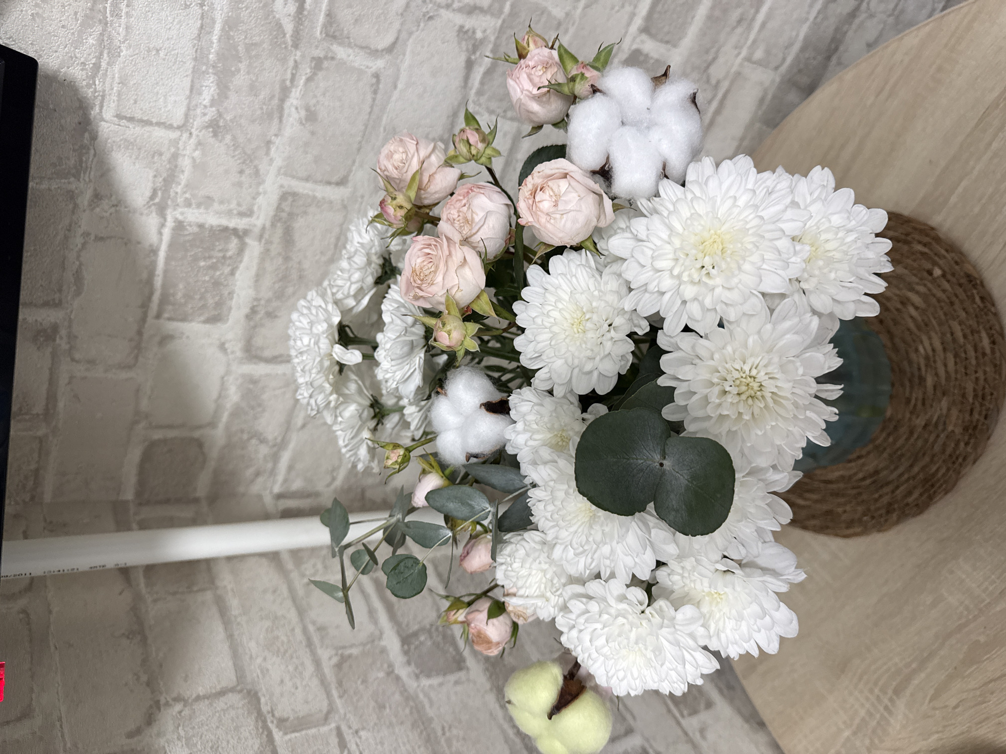 A bouquet of white chrysanthemums and pale pink roses, accented with cotton bolls, is arranged in a teal vase. The vase sits on a woven placemat on a light-colored wooden table. Behind the bouquet, a white pipe runs vertically against a light gray brick wall. The scene is indoors, likely a home or a small shop, as suggested by the brick wall texture and the tabletop. The lighting is soft, and there are no people present.