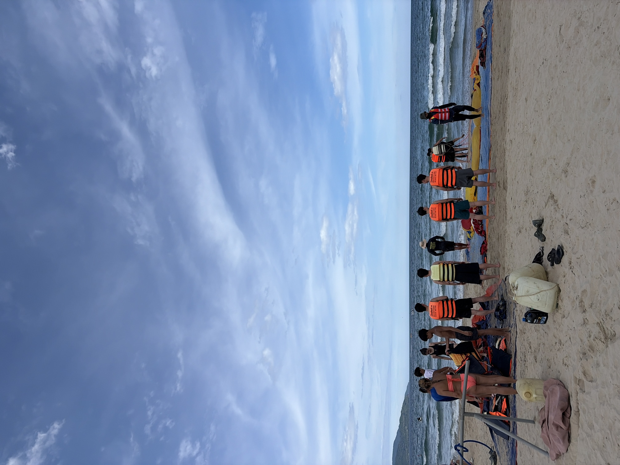 This image depicts a beach scene in Đà Nẵng, Vietnam, likely near the coast where water sports activities are offered. In the foreground, there is a sandy beach with several people gathered, some wearing life jackets and others in swimwear. Objects such as large plastic containers, folded tarps, and a pink blanket are visible on the sand. In the middle ground, the ocean stretches out with waves rolling onto the shore. A few people are standing in or near the water. In the background, a distant coastline with a green, hilly landscape is visible. The sky is partly cloudy, with patches of blue sky and white clouds. The overall atmosphere suggests a recreational setting, possibly before or after a water activity.