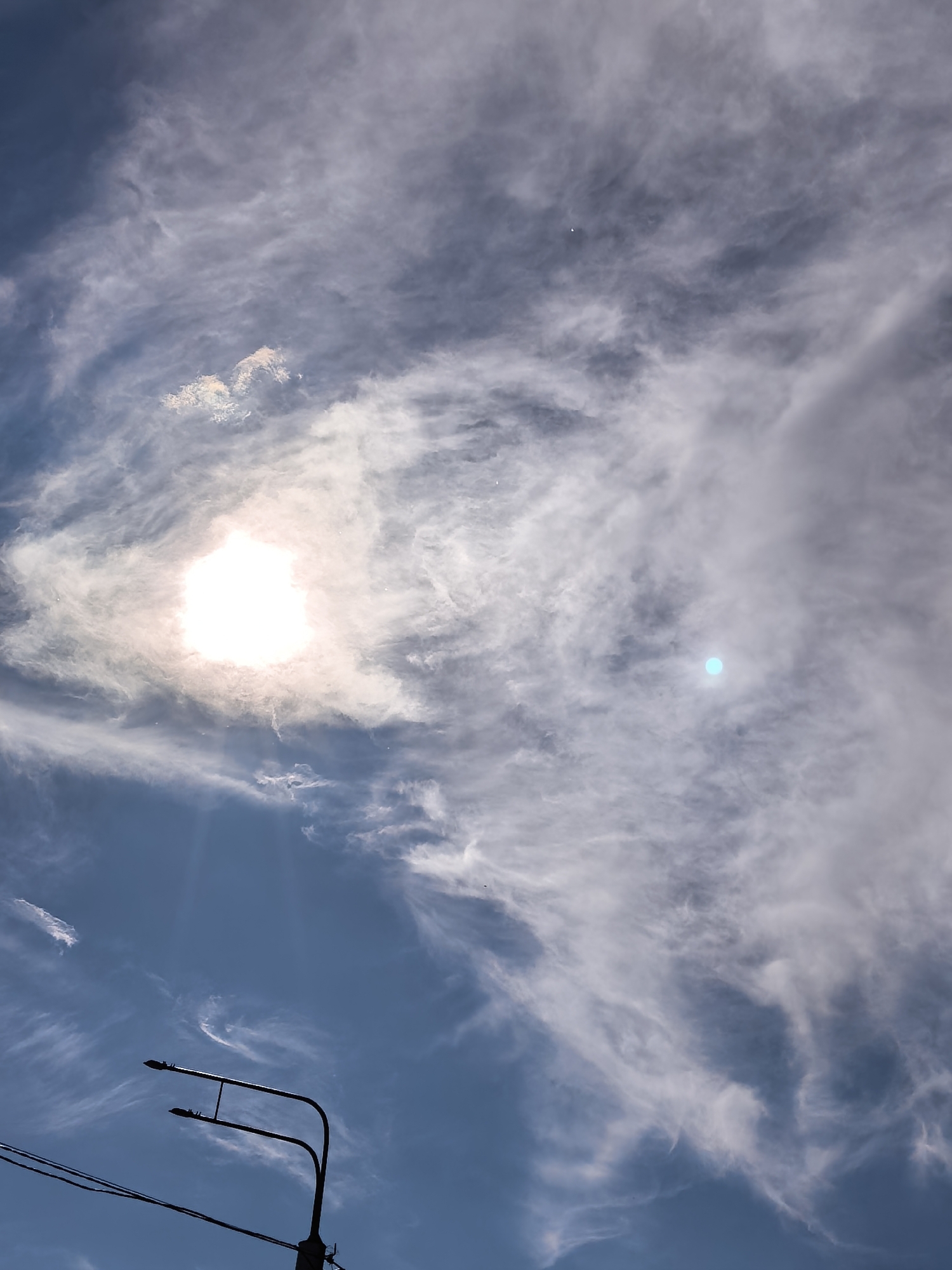 The image captures a bright, cloudy daytime sky with a dominant sun and a partial view of urban infrastructure at the bottom. The sky is filled with sweeping, wispy white and gray clouds, resembling cirrus or cirrocumulus formations, against a backdrop of deep blue.
The sun, positioned in the upper left quadrant, is an intensely bright, overexposed white circle, suggesting it is high in the sky. It is partially obscured by the clouds, creating a luminous glow and strong light rays that fan out downwards. To the right of the sun, a distinct, circular bright cyan-blue spot is visible, likely a lens flare or camera artifact rather than an object in the sky.
In the bottom left portion of the frame, the silhouette of a modern street light post with two horizontal arms is visible, along with several thin, dark power lines crossing the lower edge. These elements indicate an urban or suburban setting. No people or specific activities are depicted. The weather appears bright despite the extensive cloud cover, suggesting a clear but hazy day. No visible text is present in the image. The overall impression is one of natural light interacting with atmospheric conditions over an inhabited area.