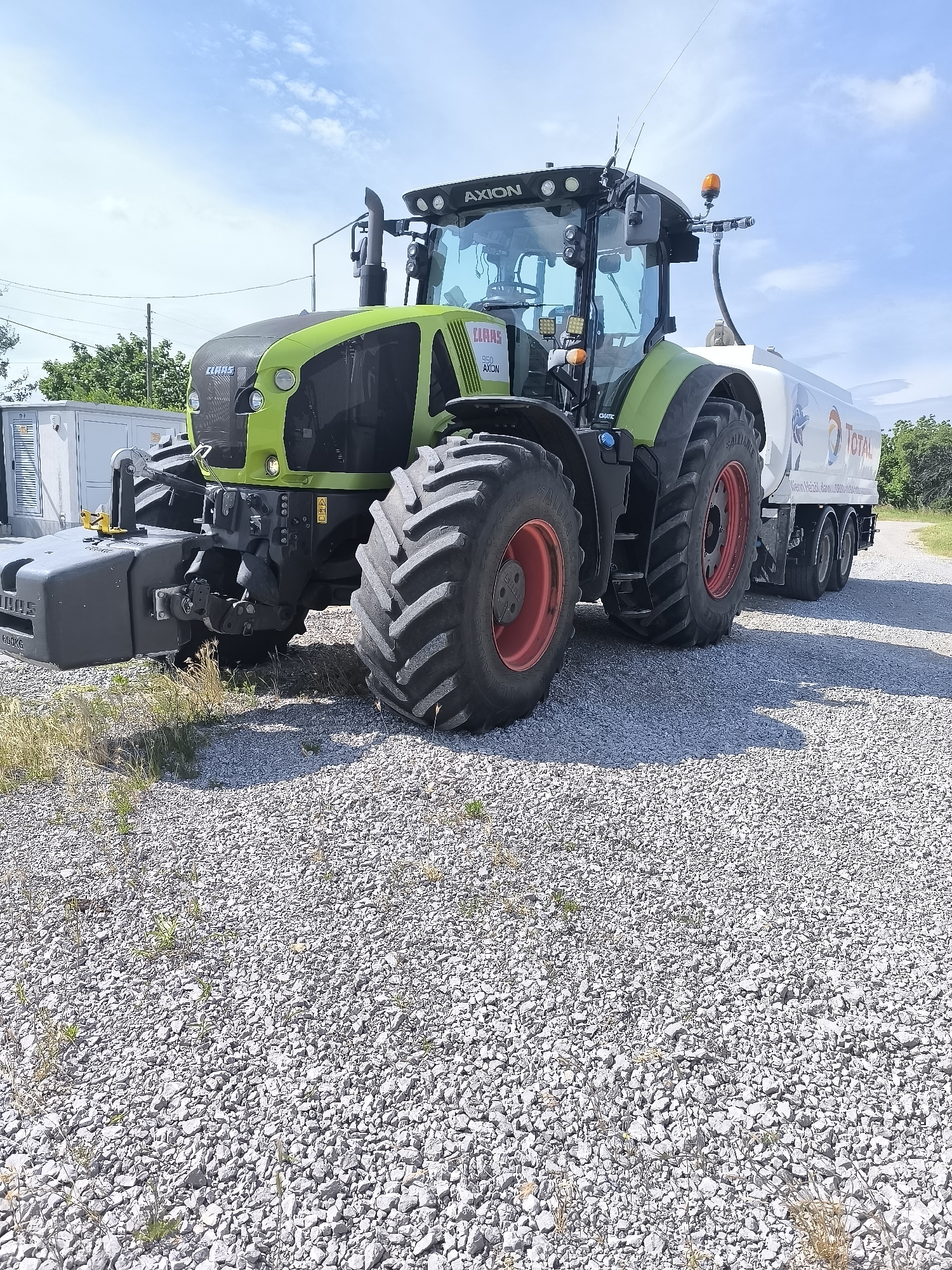 The image displays a bright, sunny day in Inovo, Bulgaria, with a large green and black CLAAS AXION tractor parked on a gravel surface. The tractor has a large white tanker trailer attached to its rear, which is branded with the "TOTAL" logo and other text. The tractor's tires are large and have red rims. In the background, there are some trees and a cloudy blue sky. To the left of the tractor, there is a white electrical box. The scene appears to be a rural or industrial area.