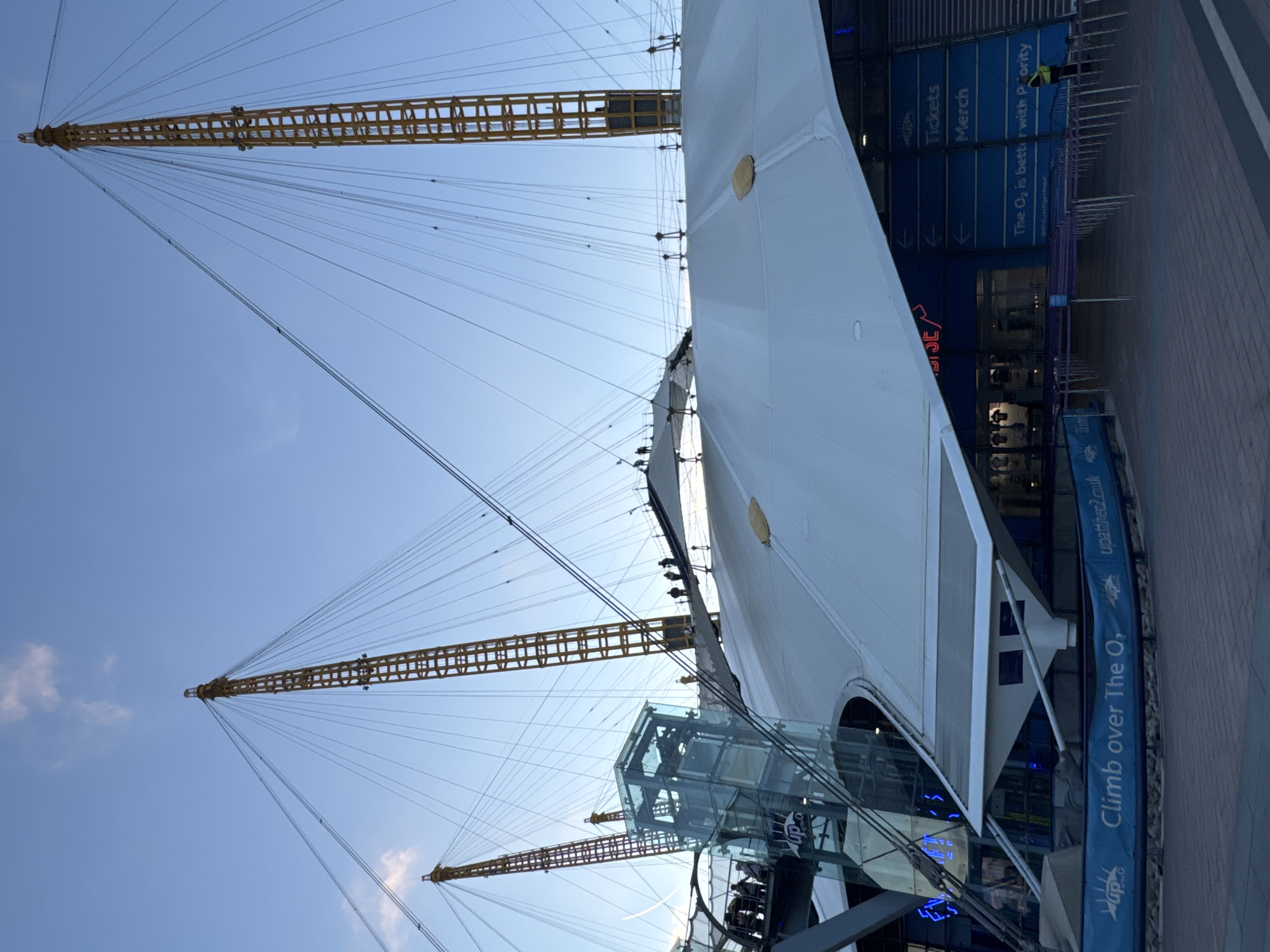 This image captures an exterior view of The O2 Arena in London, United Kingdom, under a clear blue sky with wispy clouds. The iconic structure is characterized by its large white fabric roof supported by prominent yellow, lattice-like masts. Several tension cables extend from the masts to the roof.
In the foreground, a glass elevator structure is visible, with people standing on a walkway above it. Further to the right, there are entrances to the arena with signage indicating "Tickets" and "Merch." A blue banner along the bottom of the image reads "Climb over The O₂" with a website address. A security guard in a yellow vest stands near the entrance on the right. The ground appears to be paved with brickwork. The perspective is from a low angle, looking up at the scale of the building and its supporting masts.