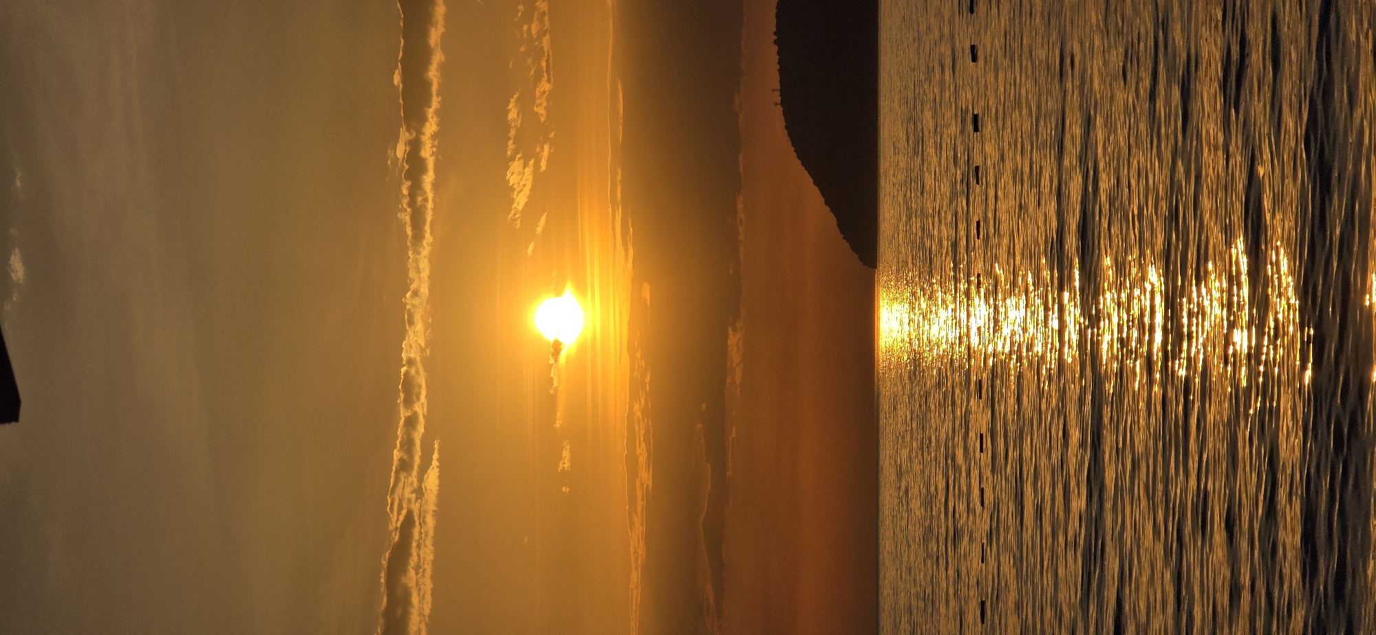 The image shows a sunset over water. The sun is the brightest object in the image and is surrounded by clouds. The water reflects the sunlight, creating a shimmering effect. On the right side of the image, there are dark objects on the water's surface, and dark land is visible on the horizon. The overall tone is warm and golden due to the sunset. This is likely located on the coastline of Bar, Montenegro, due to the coastal features present.