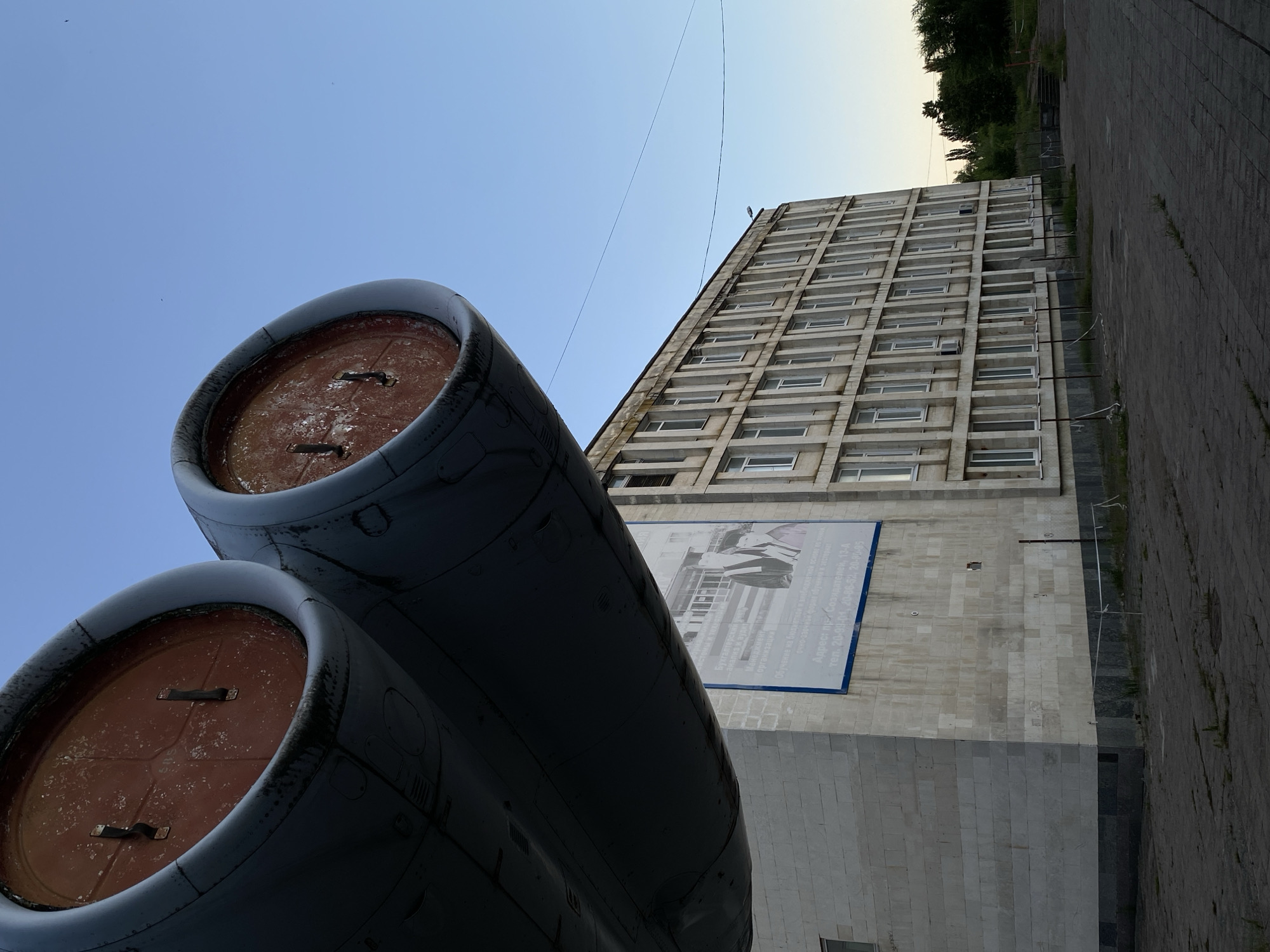 The image shows a low-angle view of two large, dark cylindrical objects in the foreground, appearing to be jet engine nacelles. Their interiors are a reddish-brown color, and they show signs of wear and tear. Behind these objects, a tall, concrete building with numerous windows stretches upwards. A large banner with a black and white image and text in Russian is affixed to the building's facade. The banner depicts what appears to be a group of people in formal attire. A paved walkway runs alongside the building on the right side of the image. The sky above is clear and a light blue, suggesting daytime. Trees are visible in the distance behind the building. The scene is likely an outdoor setting in Ulyanovsk, Russia, possibly near an aviation-related museum or monument, given the prominent display of what look like aircraft engines. The overall impression is one of weathered industrial elements and a somber architectural style.