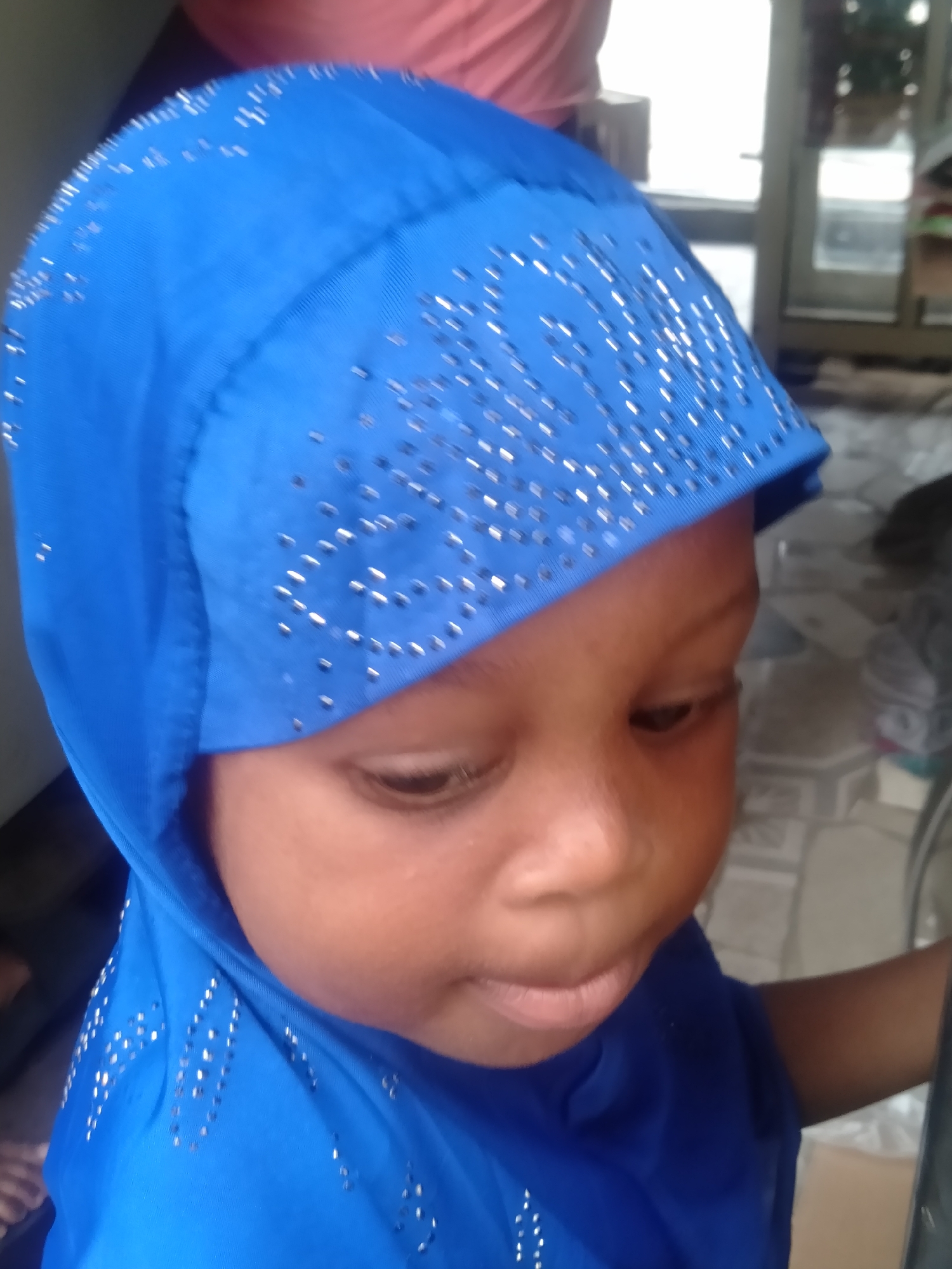 A close-up, slightly high-angle portrait features a young child, likely a toddler, with dark skin, wearing a vibrant blue head covering. The head covering, possibly a hijab, is richly embellished with numerous small, sparkling silver-colored studs or rhinestones arranged in decorative patterns across the top and sides. The child's face is central to the frame, with their head tilted slightly to the right. Their eyes are cast downwards and to the right, giving a thoughtful or observant expression. The child's mouth is slightly ajar, showing their lower lip.
The background is softly blurred, indicating an indoor setting. Indistinct shapes and colors are visible behind the child; a reddish-pink blur is seen in the upper left, while a lighter, more ambiguous area, possibly a window or doorway, appears to the right. The lighting is soft, highlighting the child's facial features and the reflective details on their headwear. The overall mood is calm and serene. The image captures a quiet moment of the child, likely in Shangisha, Nigeria. No text is visible in the image.