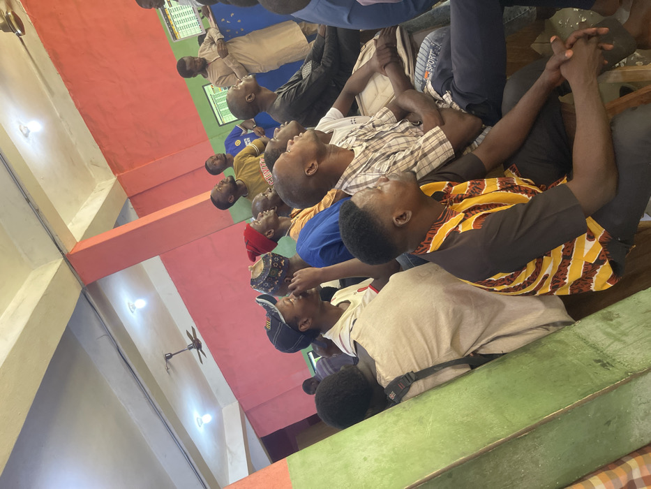 The image displays an indoor scene in Zaria, Nigeria, featuring a group of approximately twenty men, predominantly young adults. They are seated on the floor, arranged in rows, with most individuals looking upwards and towards a focal point outside the frame, suggesting attentive observation or listening. Many have their heads tilted back, and some have their hands clasped, indicating engagement.
The men wear a mix of clothing styles, from casual t-shirts and plaid shirts to more traditional attire. One man in the foreground wears a distinct brown and yellow patterned long-sleeve shirt. Another sports a blue t-shirt with "ENI" partially visible, and a cap with "NE" visible is worn by a man in the bottom right.
The interior space features walls painted in contrasting colors of red and green, alongside beige pillars. Overhead, a ceiling fan and two light fixtures provide illumination. In the background, two wall-mounted screens display text or data that is unreadable from the distance, hinting at a setting used for presentations, education, or monitoring information. The general atmosphere is one of calm attention and communal gathering, possibly for a lecture, discussion, or religious event. The bright lighting suggests it is either daytime indoors or a well-lit evening event.