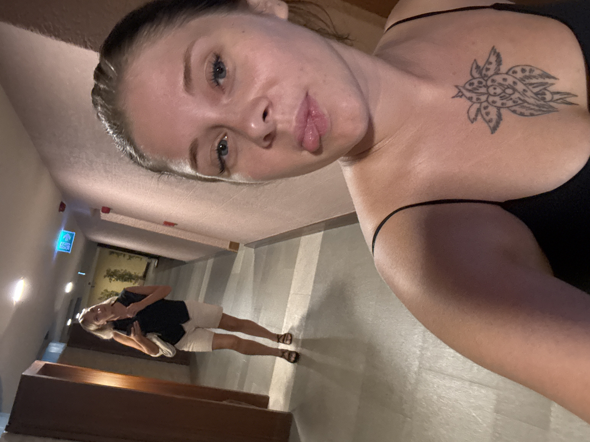 A selfie is taken by a woman in the foreground, who is leaning back slightly and sticking her tongue out. She is wearing a black spaghetti strap top and has a tattoo on her chest. Behind her, another woman is walking down a hotel corridor. She is wearing a black vest, white shorts, and sandals, and is holding a phone in her hand. The corridor has beige walls and a tiled floor, with wooden doors and potted plants visible. An illuminated exit sign is mounted on the ceiling. The location appears to be a hotel in Aksu, Turkey, likely within the resort area.