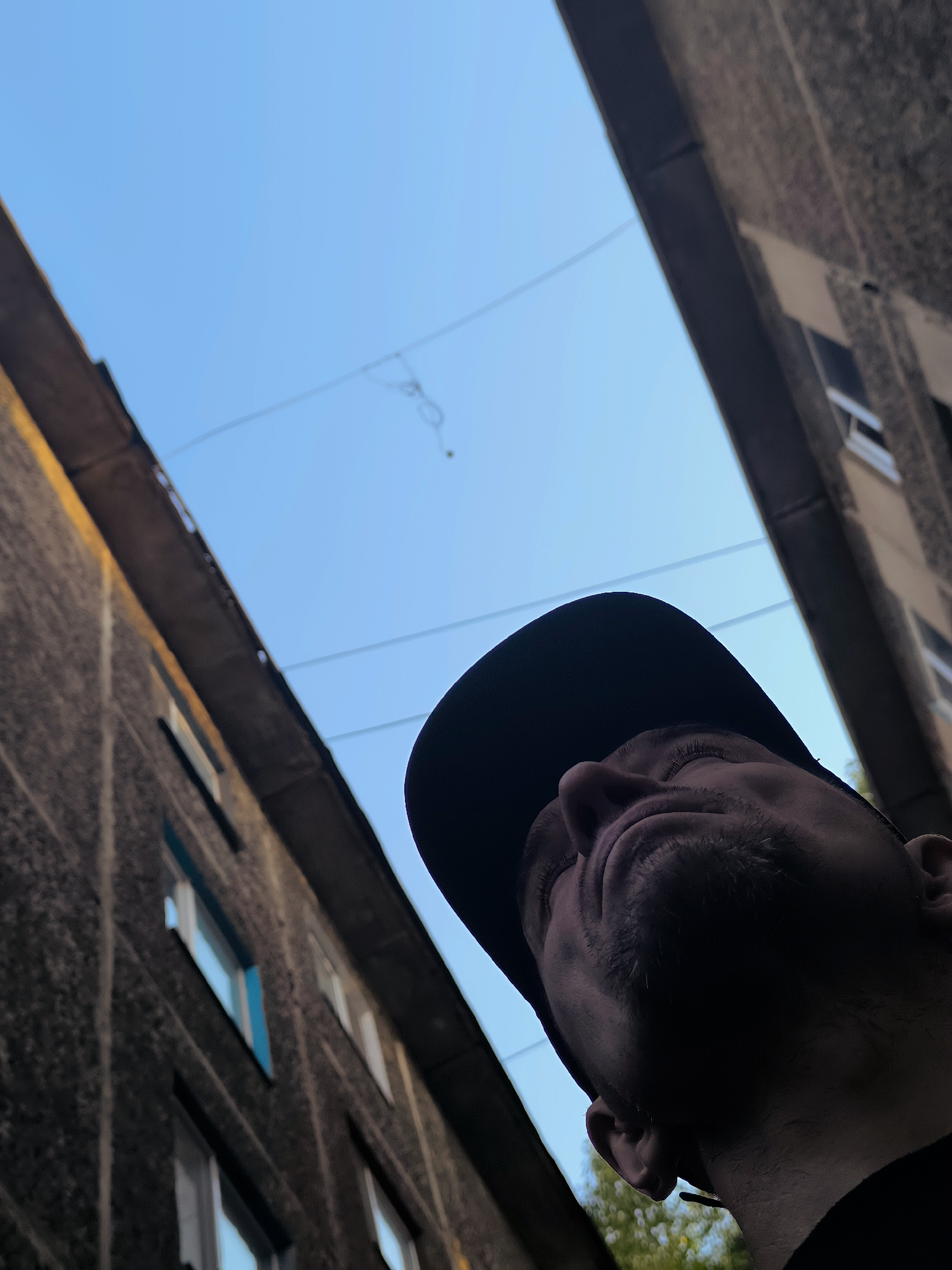 The image shows a close-up, low-angle view of a man's head and shoulders, wearing a black baseball cap. He is looking upwards, and his face is partially obscured by shadow and the cap. The background is a clear blue sky, with two buildings visible on either side, creating a narrow passage. The buildings appear to be made of concrete and have several windows. There are power lines strung across the sky. The image seems to be taken in an urban setting, possibly in Qarağandı city administration, Kazakhstan, though this cannot be definitively confirmed from the visual information alone. The lighting suggests it is daytime.