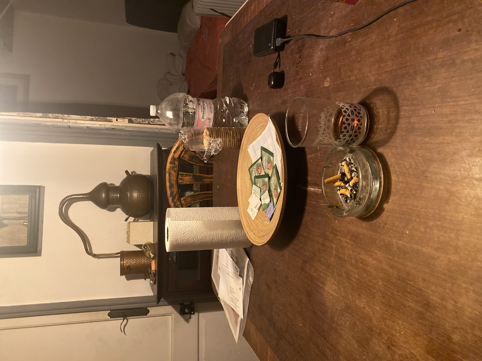 The image depicts an indoor scene of a table in what appears to be a residence or perhaps a dimly lit establishment. The table is made of wood and shows signs of wear. On the table, there are several objects. In the foreground, there is a glass ashtray containing cigarette butts. Next to it is a glass with amber liquid and a distinctive patterned design on its exterior.
Further back on the table, a wooden bowl holds a collection of small packets, possibly tea or medication, along with some papers. A roll of paper towels stands upright beside the bowl. To the right of the bowl is a clear plastic bottle of "Fuente Primavera" water. Adjacent to the water bottle, there is a black power bank plugged into a black cable, and a small black device, possibly earbuds, rests nearby. Behind these items, a stack of crackers in plastic packaging is visible.
In the background, against a cream-colored wall, a dark wooden cabinet is present. On top of the cabinet, there is a decorative copper distillation apparatus with a round base and a curved pipe. Next to it is a small, textured copper container, and a white square object, possibly a small box or a paper item. A framed picture hangs on the wall above the cabinet. A wooden chair with a rustic design is partially visible to the right of the cabinet, and beyond that, a glimpse of a bed with orange and white bedding can be seen. A closed white door with a dark handle is visible on the left side of the image.
The overall lighting is dim and warm, suggesting evening or nighttime. The presence of the distillation apparatus and the general ambiance might suggest a bohemian or artistic dwelling, but there are no explicit indicators of its location. Without additional context or information, it is impossible to definitively determine if this scene is located in the city of Barcelona, Spain.