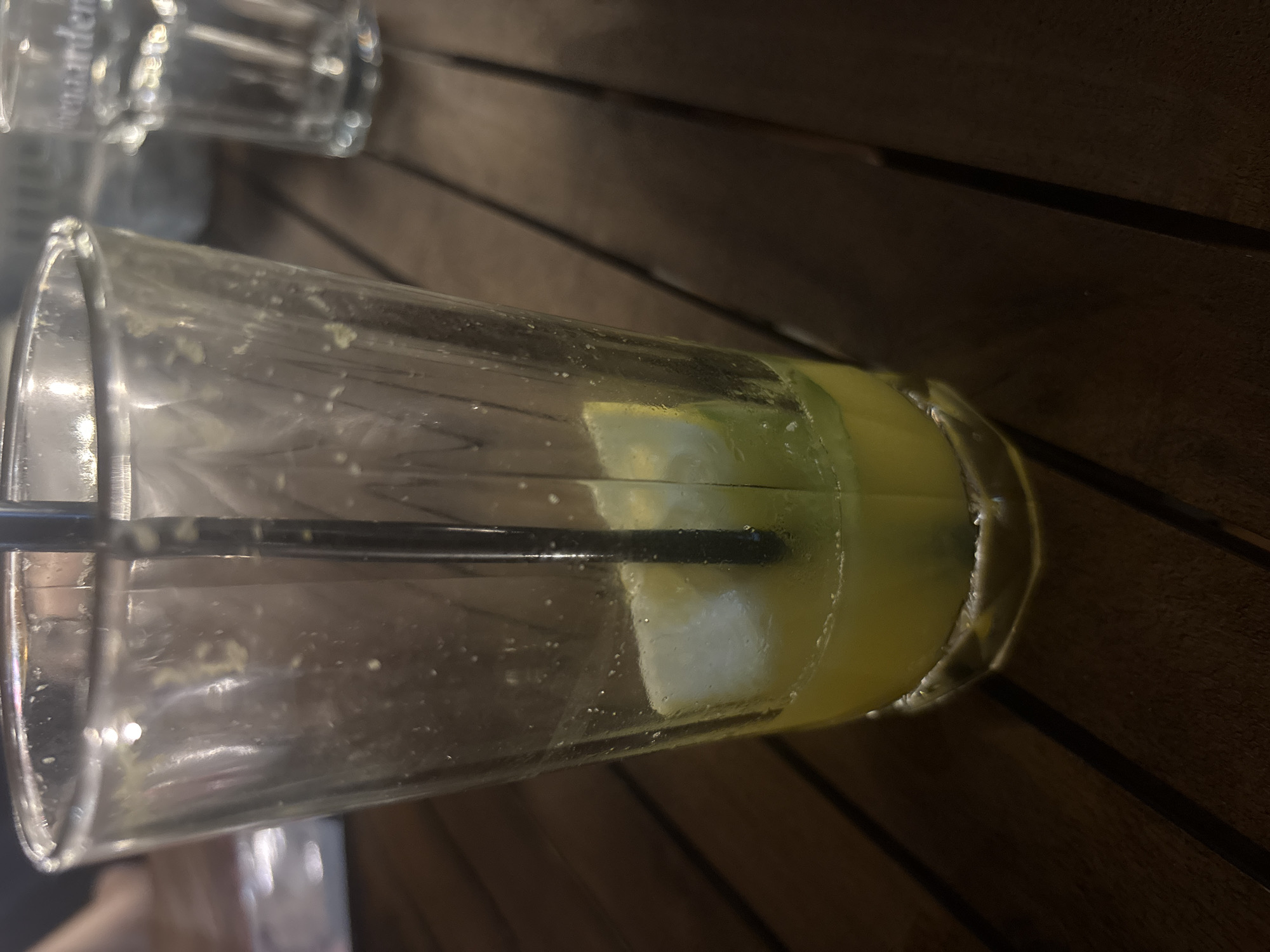 The image shows an empty, tilted glass on a dark brown wooden table, likely outdoors. The glass contains remnants of a yellow drink, possibly lemonade or a similar citrus beverage, with a straw inside. A slice of lemon or lime is visible at the bottom of the glass. Another glass, partially out of focus, is visible in the top left corner of the image. The scene suggests a casual setting, potentially in a restaurant or bar in Plovdiv, Bulgaria, although without further context, the exact location is impossible to determine. There are no people in the image.