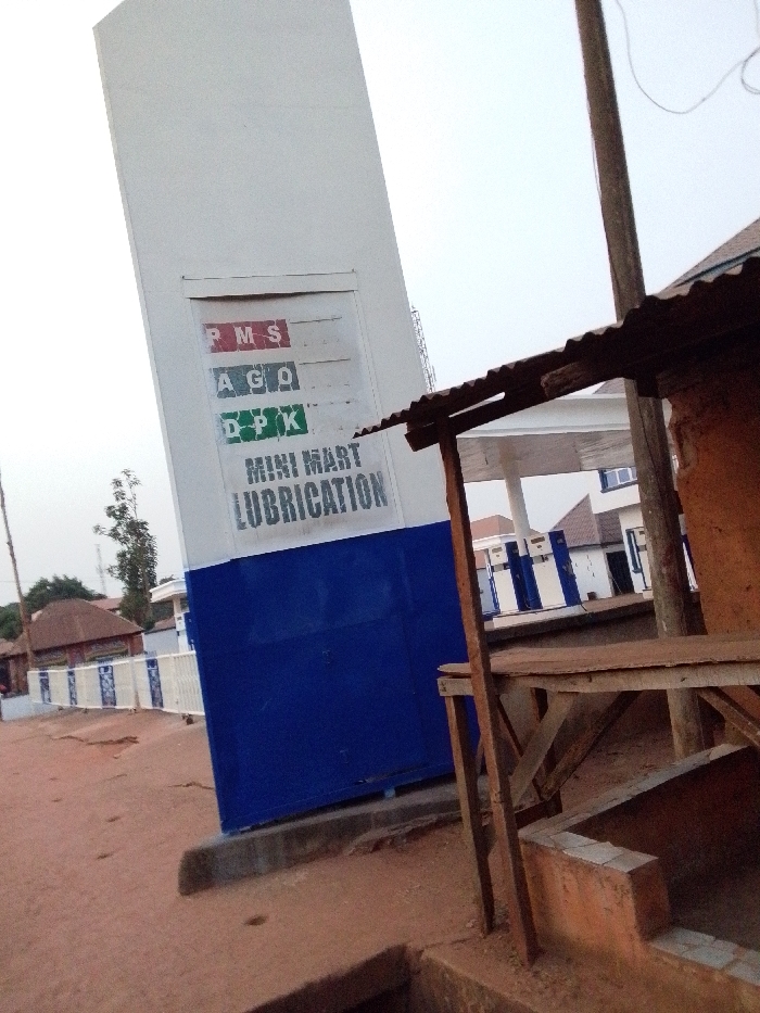 This image depicts a petroleum fueling station located in Ankpa, Nigeria. In the foreground, on the left, a tall, white and blue rectangular sign is visible. The white upper section of the sign features three labeled rectangular panels for fuel types: "PMS" (red letters), "AGO" (green letters), and "DPK" (green letters). Each panel has an empty slot for price display, indicated by a dollar sign next to "PMS." Below these, the words "MINI MART" and "LUBRICATION" are stenciled in black. The lower half of the sign is solid blue.
To the right of the sign, partially obscuring the view, is a rustic, makeshift structure. This structure consists of wooden posts supporting a corrugated metal roof and a rough wooden shelf. Below this shelf, a small, unrendered brick or concrete structure with a square opening is present at ground level.
The ground in the immediate foreground and extending to the left appears to be red-brown earth or dirt, with some unevenness and shallow depressions. Behind the main sign, a low white fence with blue accents runs horizontally, separating the station area from what appears to be a dusty road. In the background, beyond the fence and a tree, several single-story buildings with reddish roofs are visible, consistent with local architecture. To the far right, part of the main fueling station canopy, supported by white and blue striped pillars, can be seen. A wooden utility pole with wires runs vertically on the right side of the image, near the makeshift structure. The sky is overcast and light. No people or vehicles are visible.