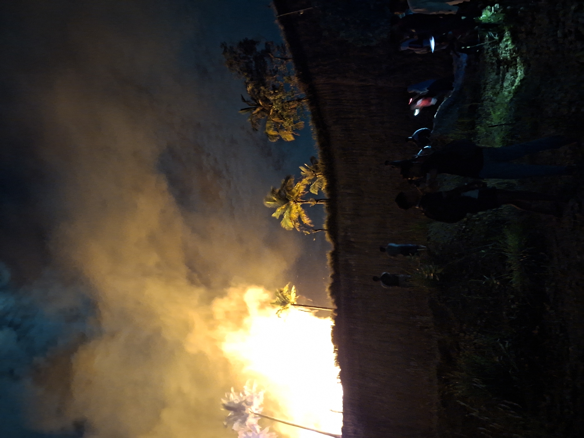 At dusk, in Lhokseumawe, Indonesia, a field is burning, with large flames and smoke obscuring the sky near palm trees. A group of people are watching from the edge of the field.
