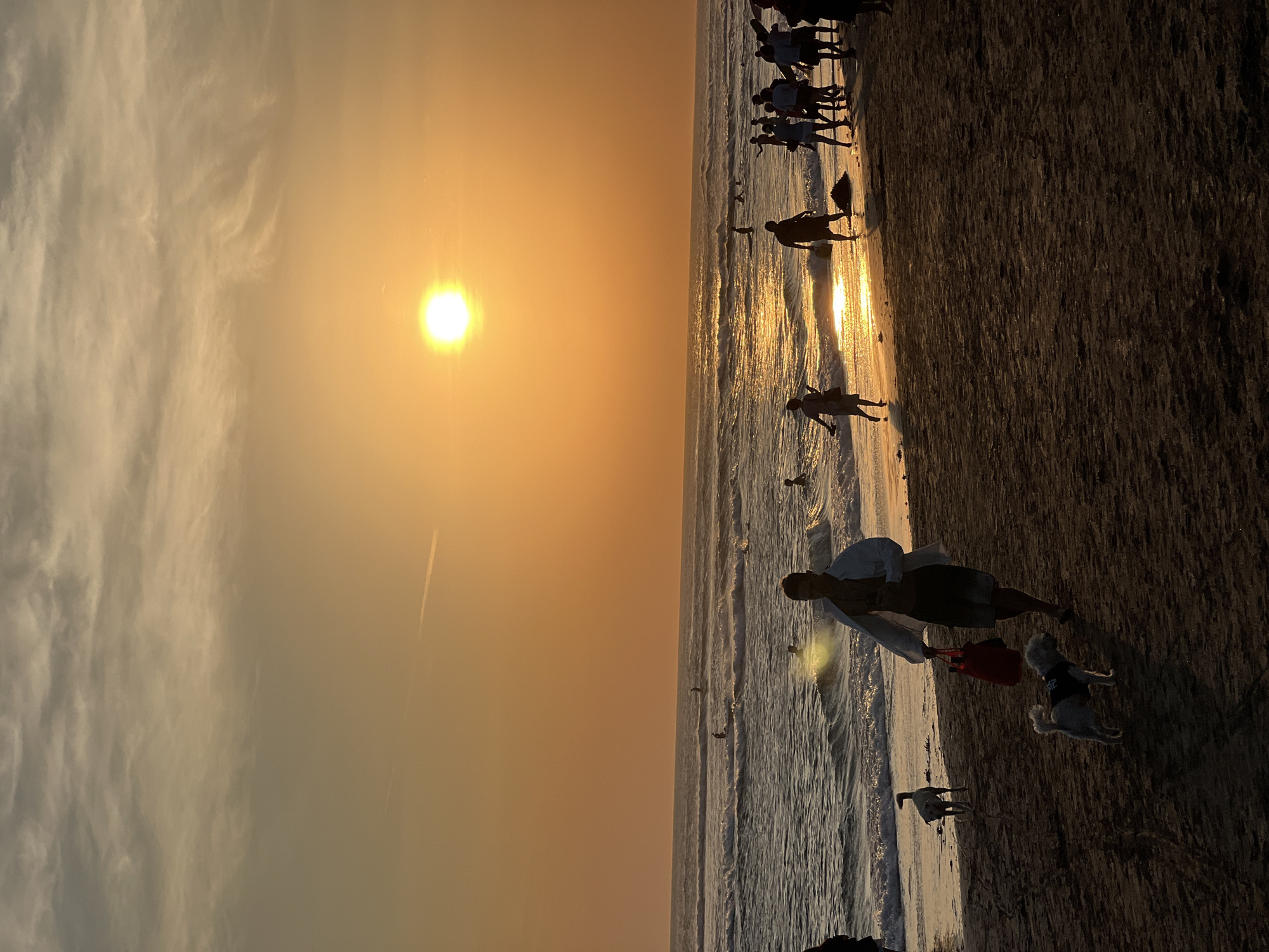 At sunset on a beach in North Kuta, Indonesia, a group of people are visible. The sun, a bright orb, is low on the horizon, casting a warm orange glow over the sky and water. Gentle waves roll onto the sandy shore, and several individuals are wading or surfing in the ocean. In the foreground, a man walks with two dogs, one small and white wearing a dark vest, and another a lighter color. The man carries a red bag. Other people are scattered along the beach, some in groups and some individually, enjoying the evening. The overall scene is peaceful and depicts a typical sunset on a popular beach.
