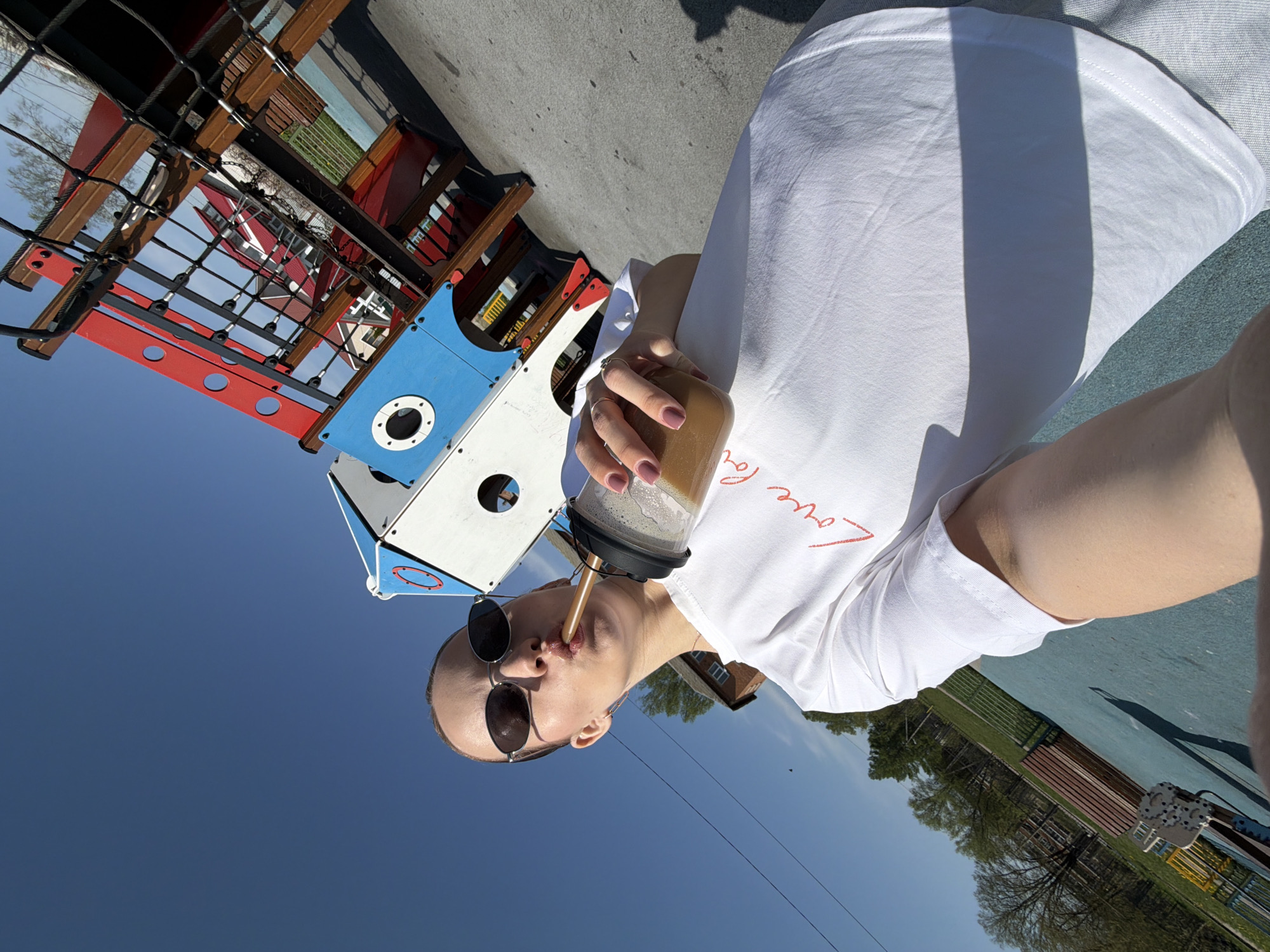 A person wearing sunglasses and a white t-shirt is drinking from a clear plastic cup with a straw while standing in a playground in Reshetnikovo, Russia. The playground features a climbing structure with red, blue, and white elements, including a rope net. The sky is clear and blue. The background shows trees and buildings, and the playground surface appears to be a blue rubber material.